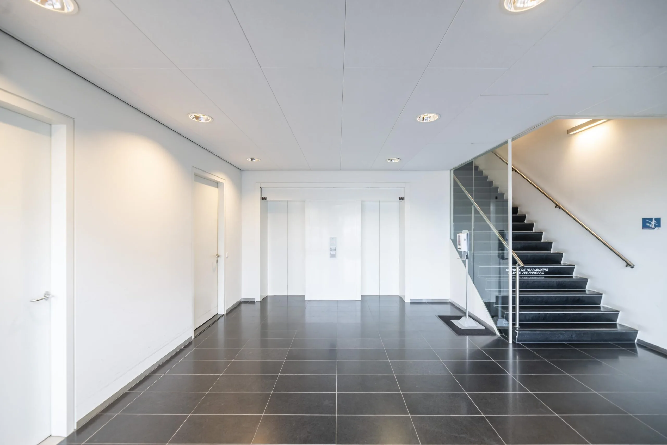 Moderne entreehal met zwarte tegelvloer, witte wanden, een liftdeur in het midden en een trap met glazen leuning rechts.