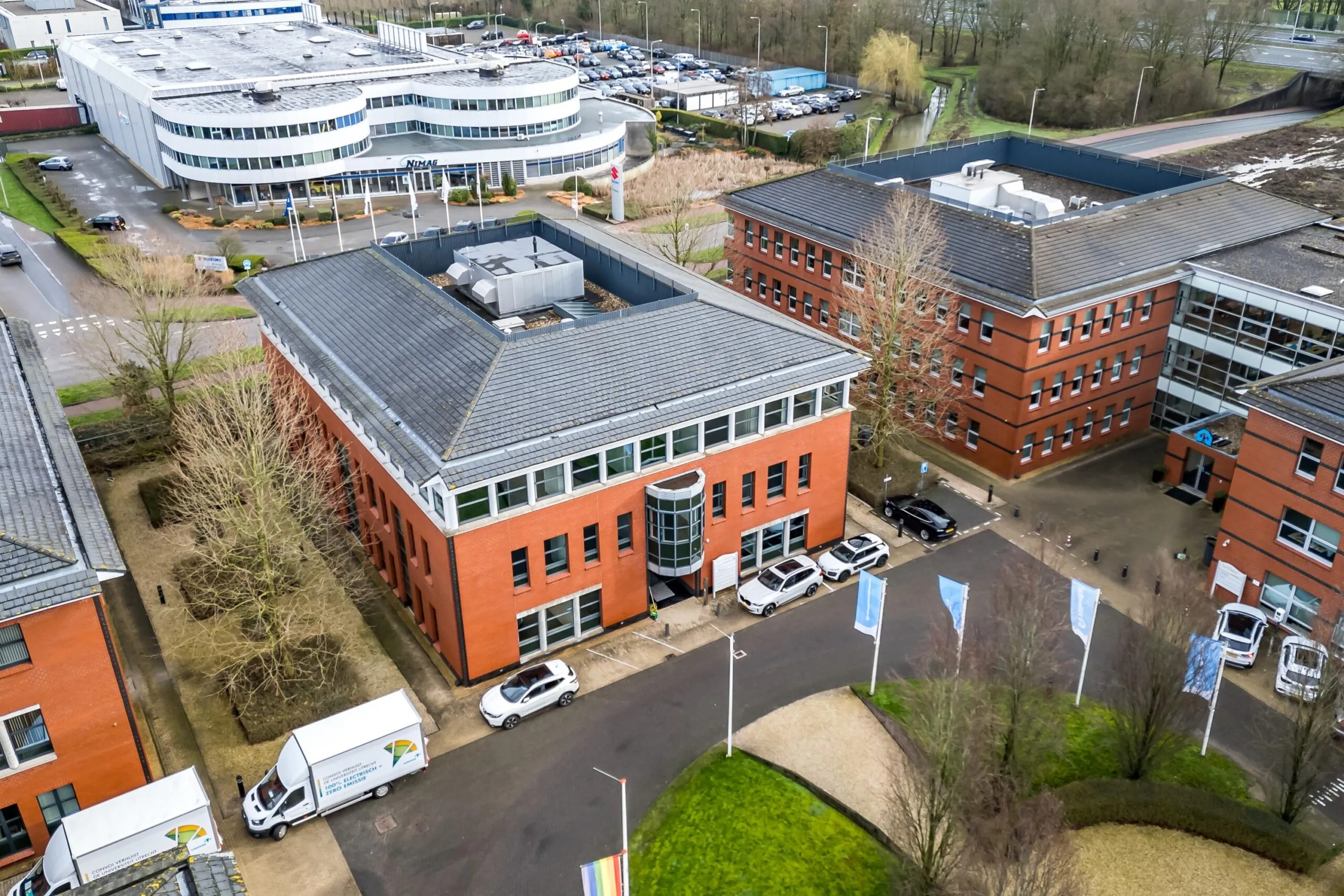 Luchtfoto van een modern kantoorgebouw aan de Lange Dreef in Rijswijk met omliggende parkeerterreinen en bedrijfswagens.