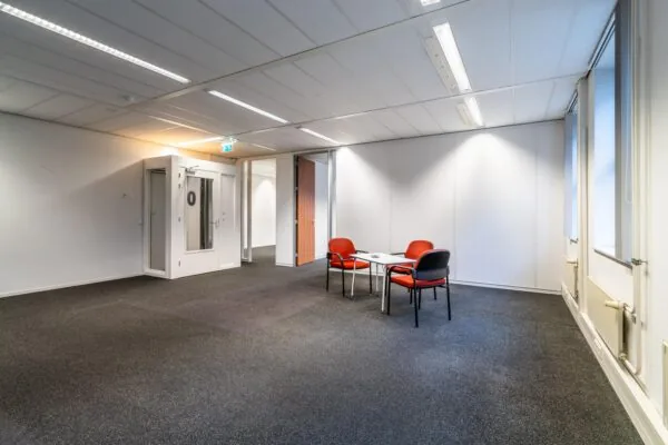 Kantoorruimte aan de Lange Dreef met tapijtvloer, witte wanden en een zithoek met drie rode stoelen rond een tafel.