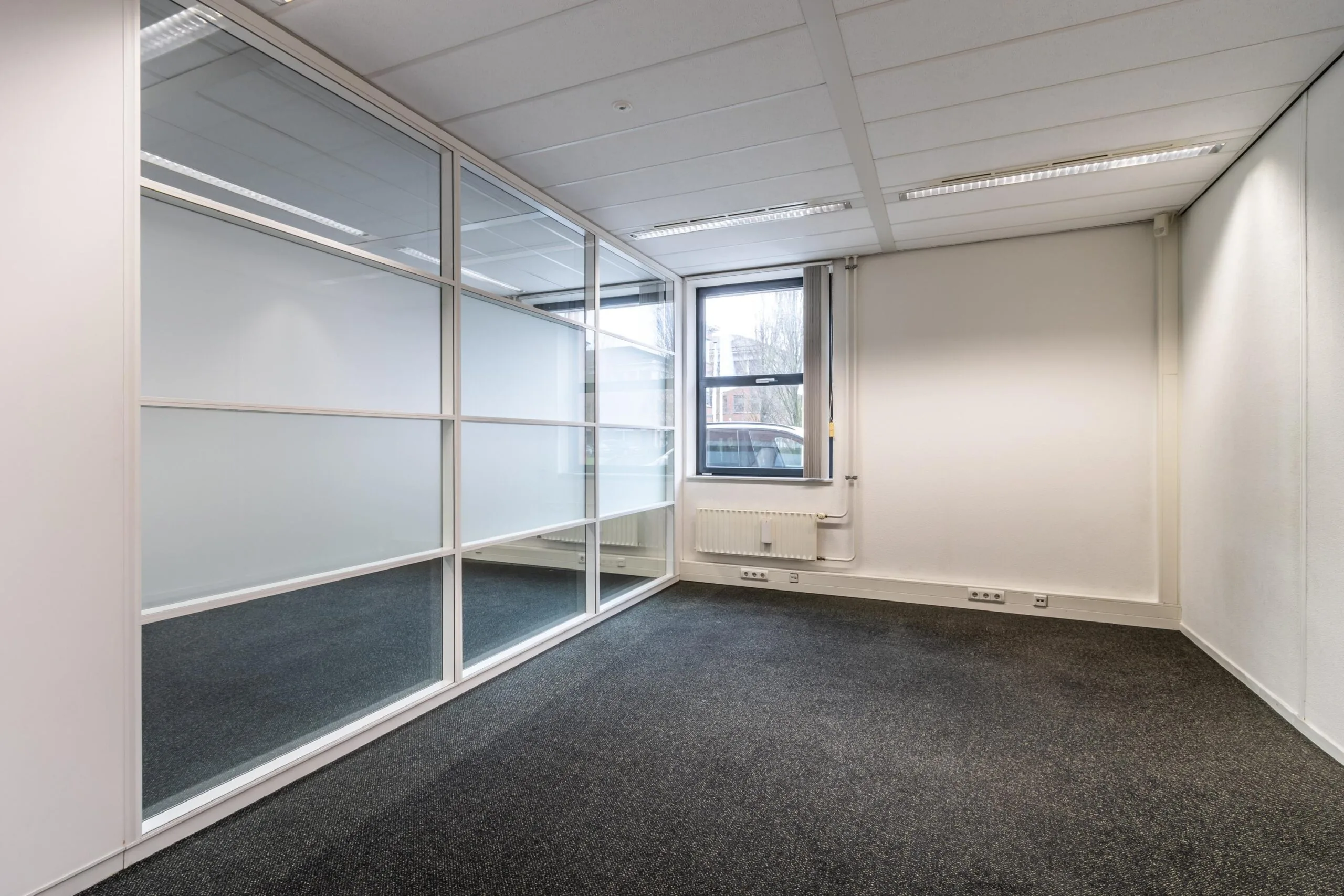 Leeg kantoor met tapijtvloer, systeemplafond, glazen scheidingswand en een raam aan de Lange Dreef.