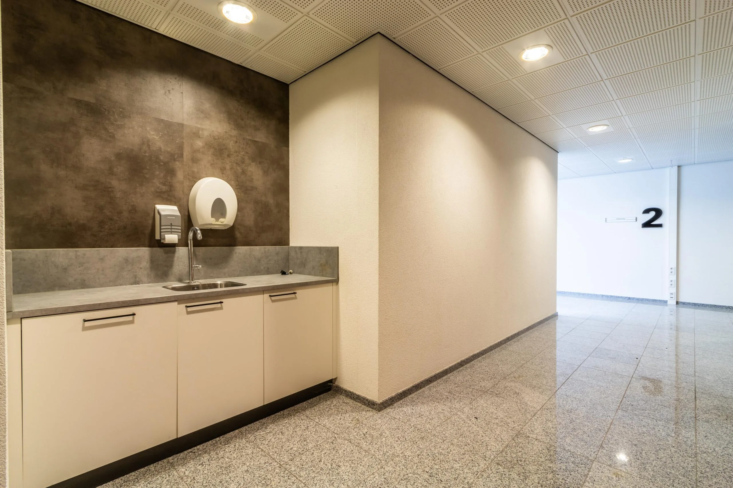 Keukenblok met gootsteen en zeepdispensers in een strakke, moderne gang op verdieping 2 van een kantoor- of bedrijfsgebouw aan de Lange Dreef.