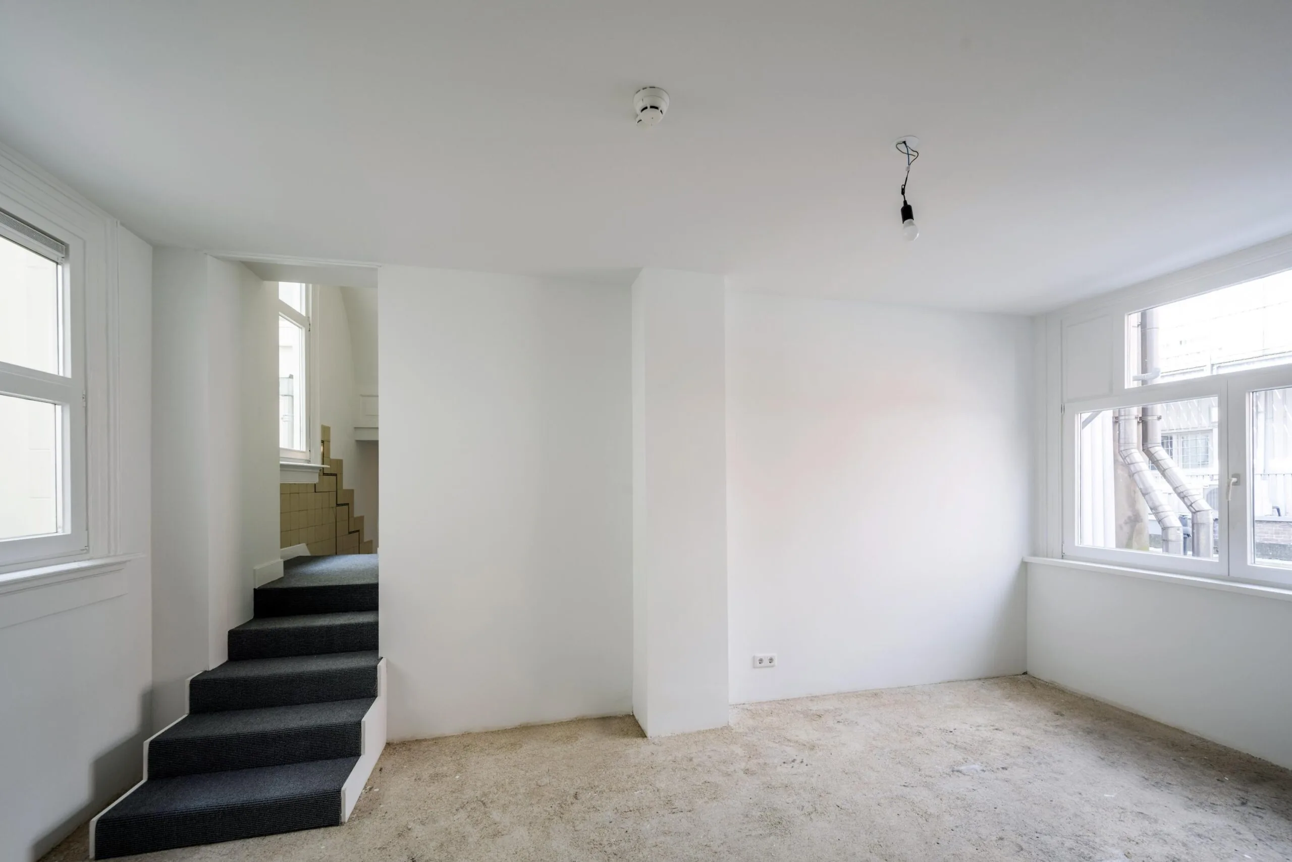 Lege, witte kamer aan het Damrak met onafgewerkte vloer, grote ramen en een trap naar een hoger gelegen deel.