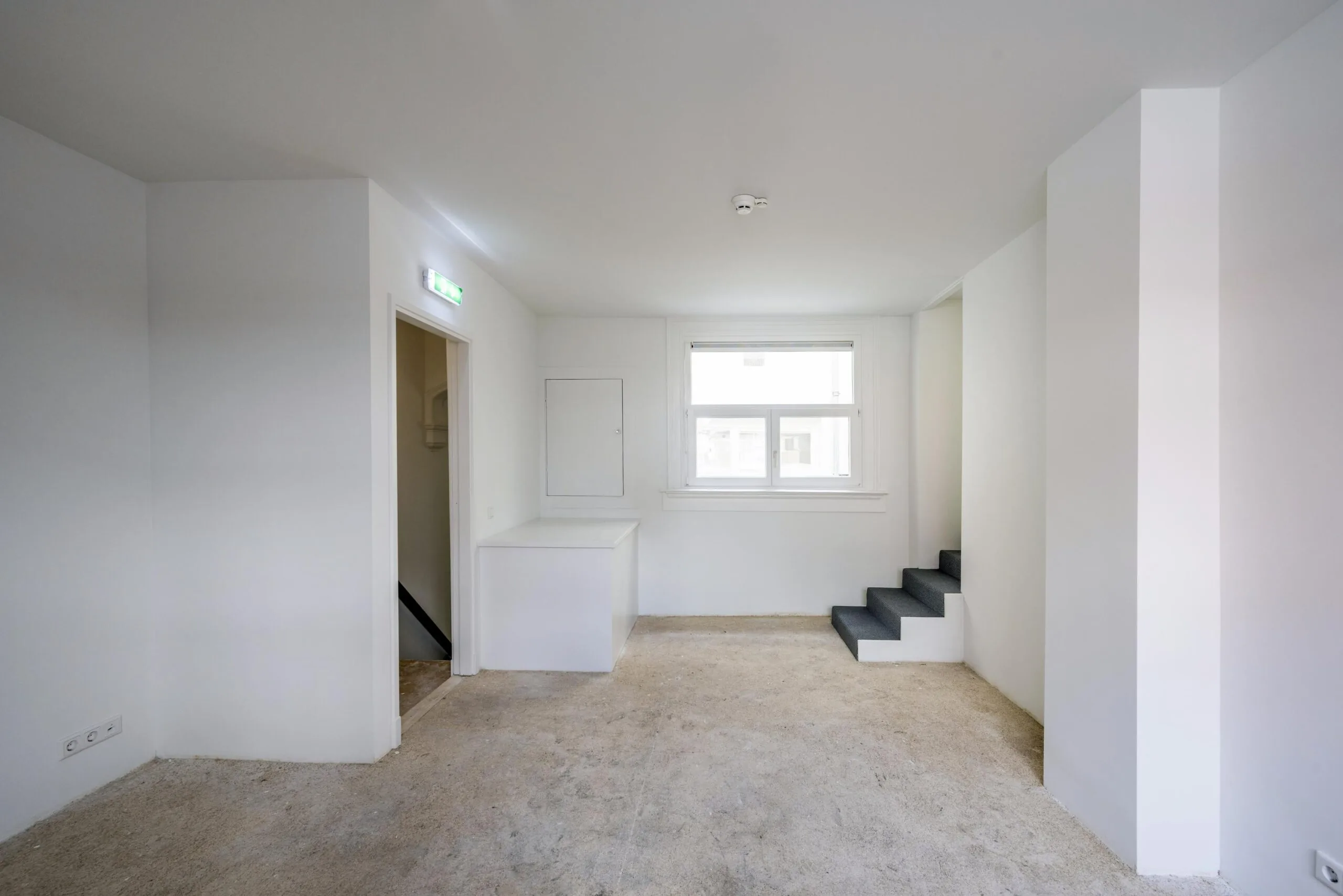 Leeg, wit interieur met betonnen vloer, een raam, kleine trap en deuropening in een ruimte aan het Damrak.