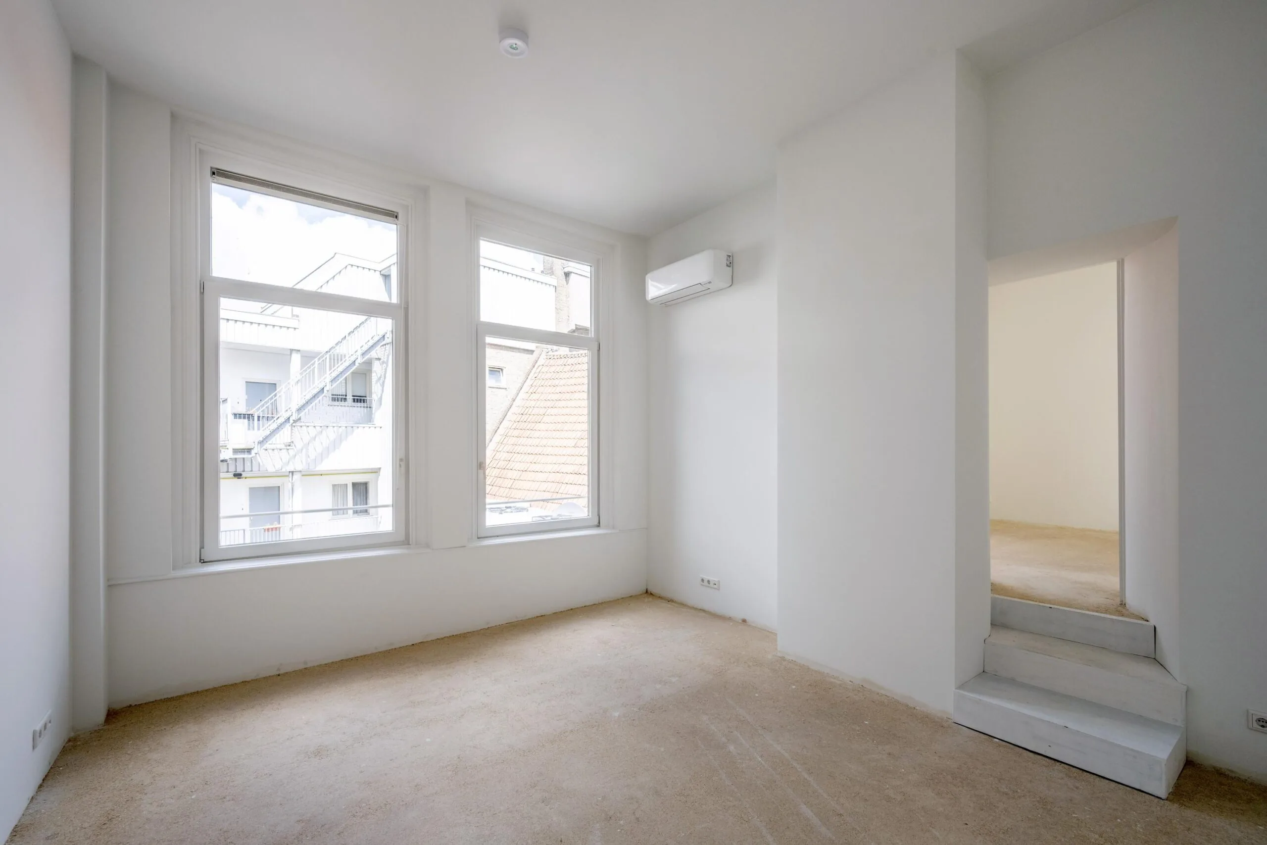 Lichte, lege kamer in een appartement aan de Damrak met grote ramen, witte muren en een doorgang naar een aangrenzende ruimte.