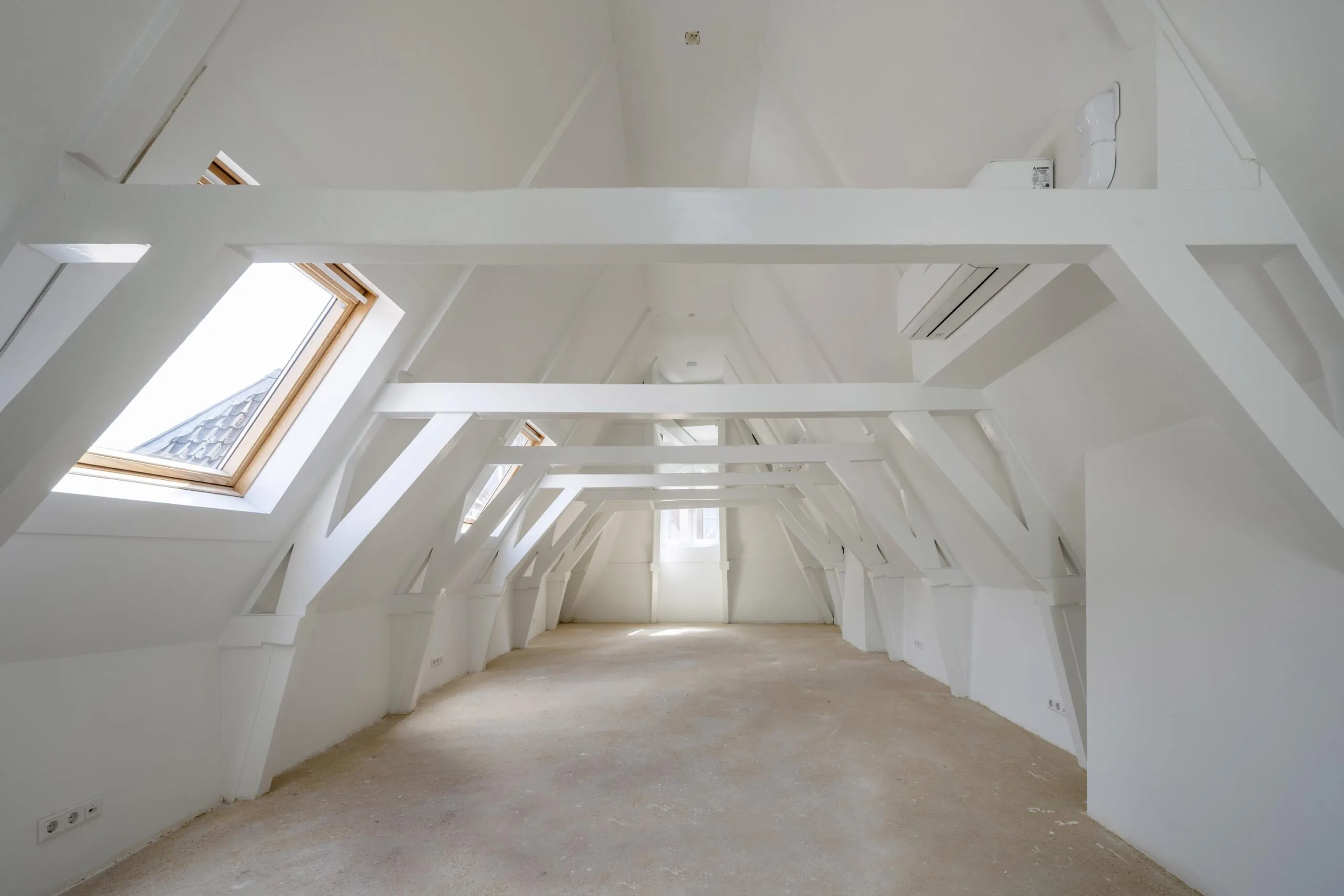 Lichte, leegstaande zolderruimte met witgeschilderde houten balken en dakramen aan het Damrak.