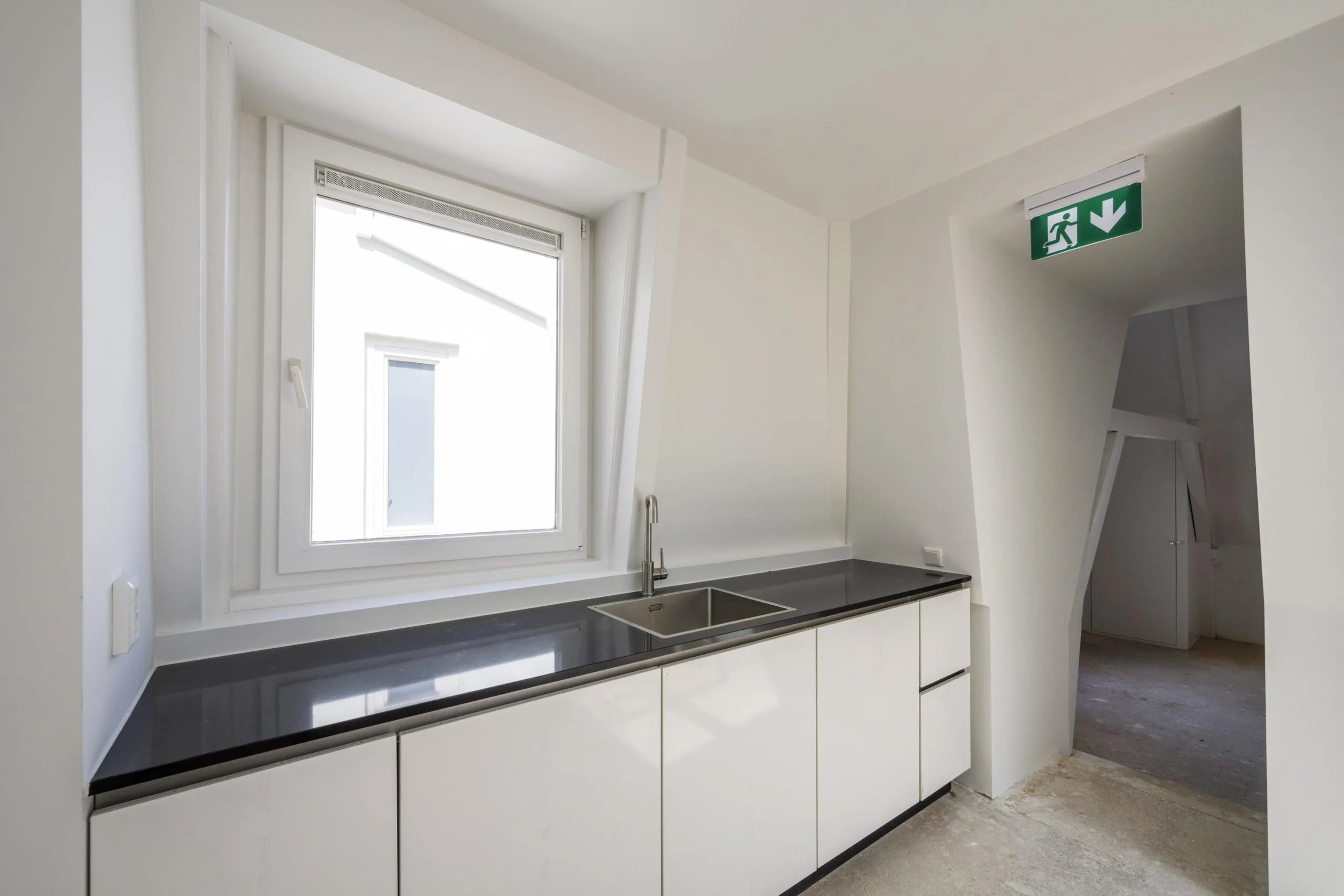 Minimalistische witte kitchenette met zwart aanrechtblad bij een raam op de Damrak, met nooduitgangsbord zichtbaar.