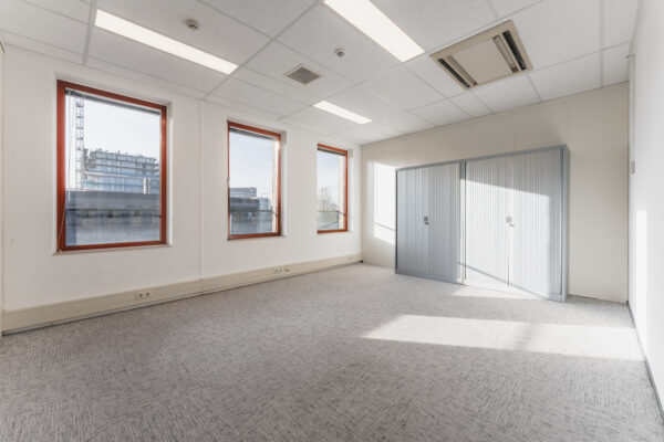 Lege kantoorruimte met tapijtvloer, drie ramen en een metalen archiefkast.