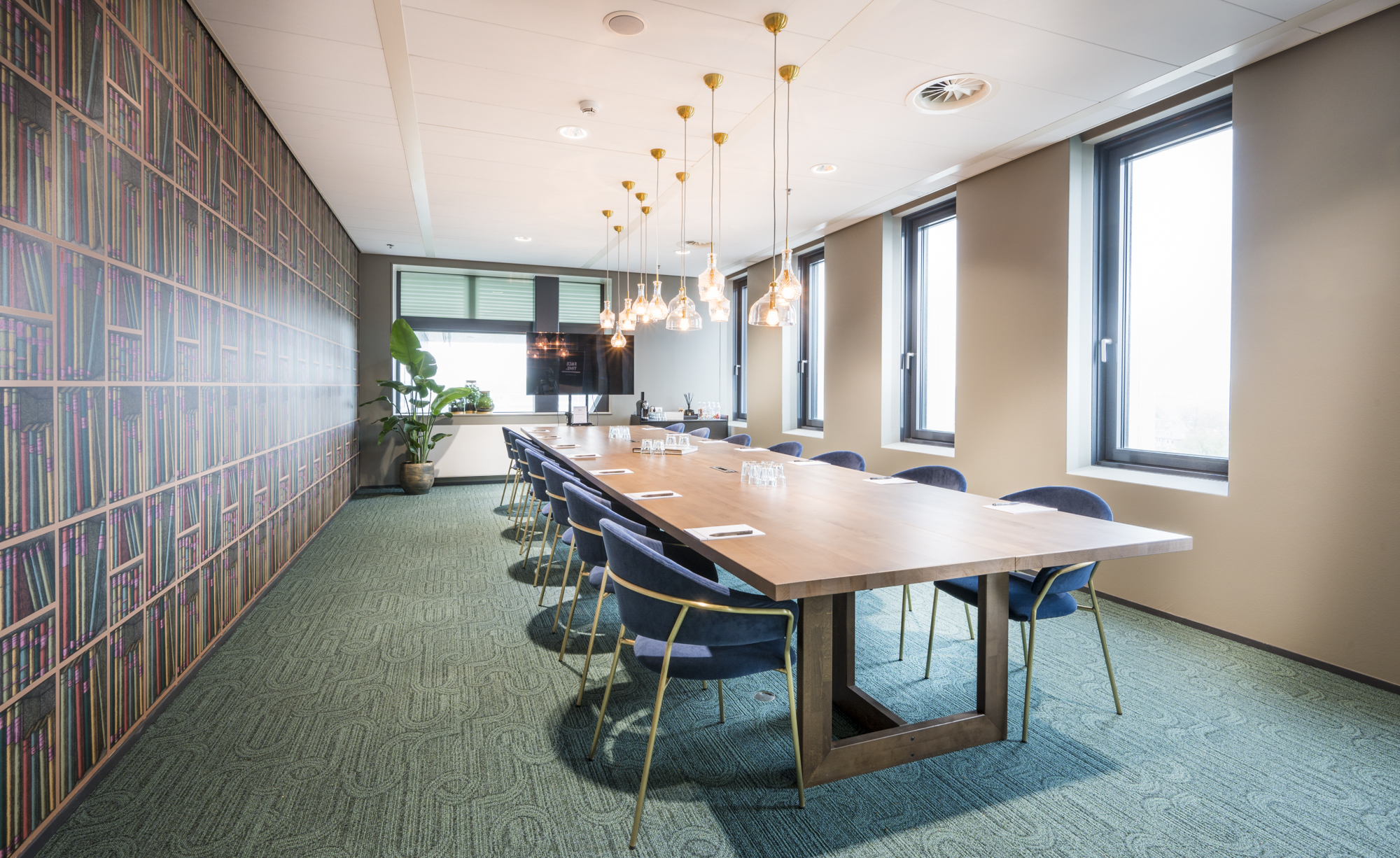 Vergaderruimte met een lange houten tafel, blauwe stoelen, een boekmuur op de achtergrond en moderne hanglampen aan het plafond.