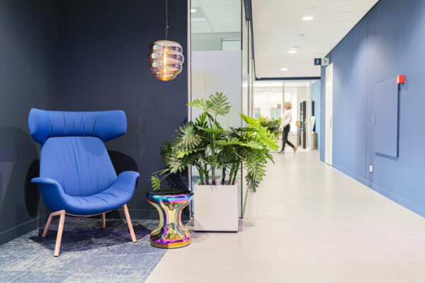 Een moderne kantoorruimte met een blauwe stoel, een kleurrijk krukje, en een groene plant naast een gang.