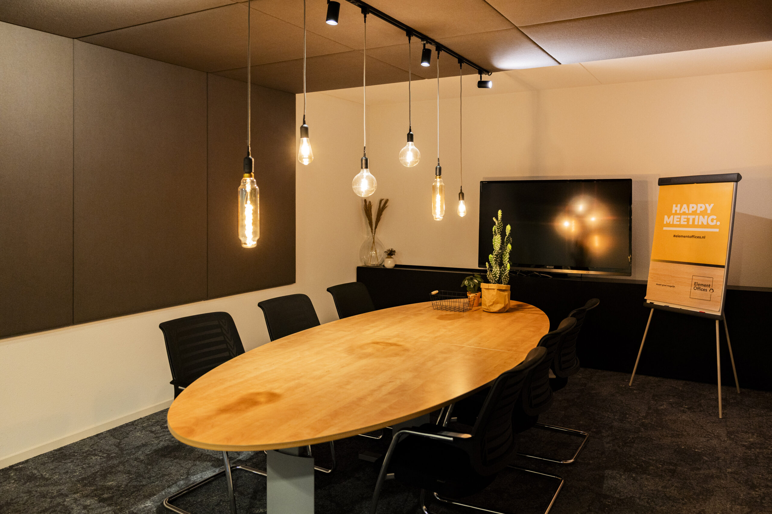 Een moderne vergaderruimte met een houten tafel, stoelen, hangende lampen, een scherm en een flip-over met de tekst "HAPPY MEETING" in een professioneel kantoorinterieur.