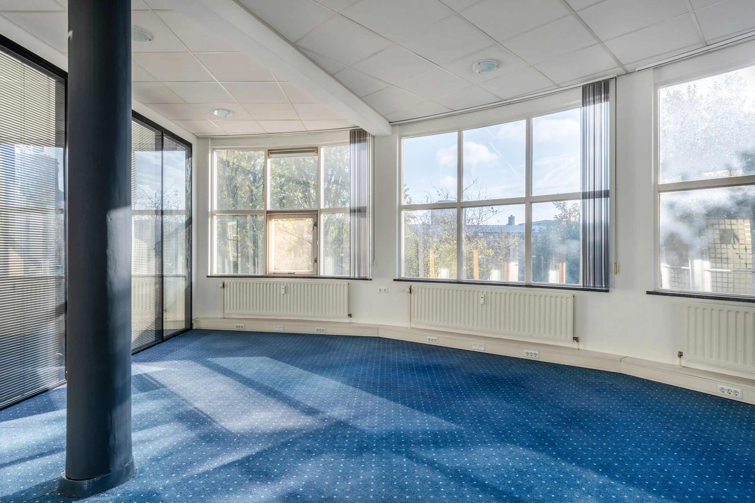 Bright, curved office space on Rozenlaan with large windows, blue carpet, and vertical blinds.