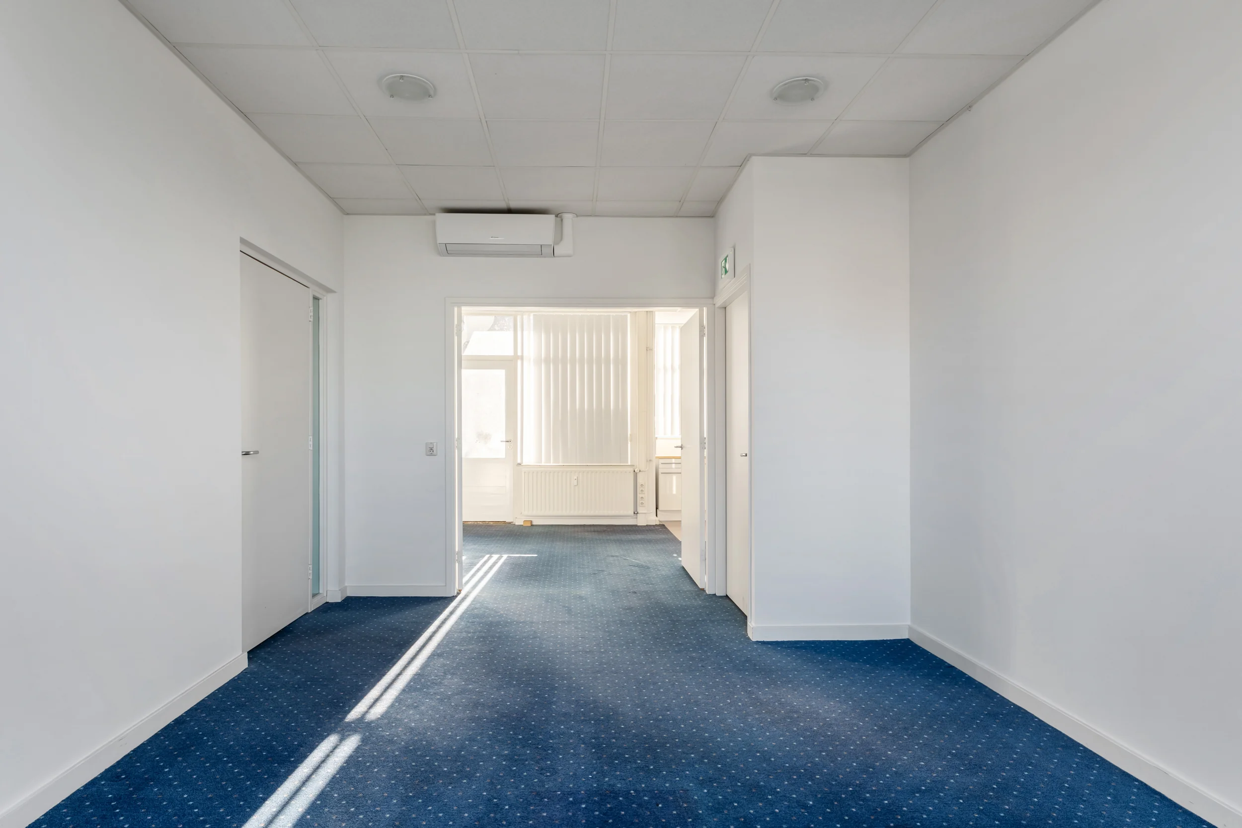 Bright hallway in a building on Rozenlaan with blue carpet, white walls, and a visible air conditioning unit.