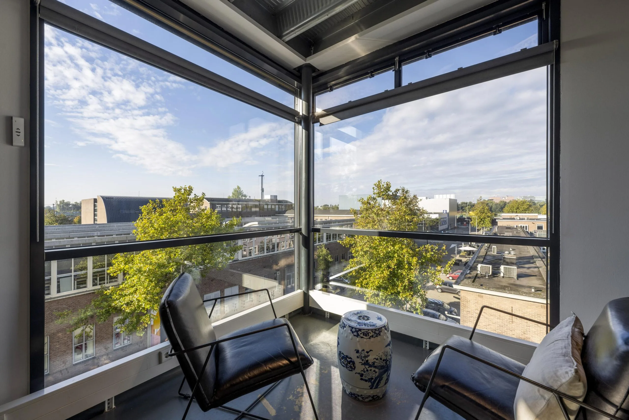 Corner seating area with two black chairs, a decorative ceramic stool, and large windows overlooking Helicopterstraat.