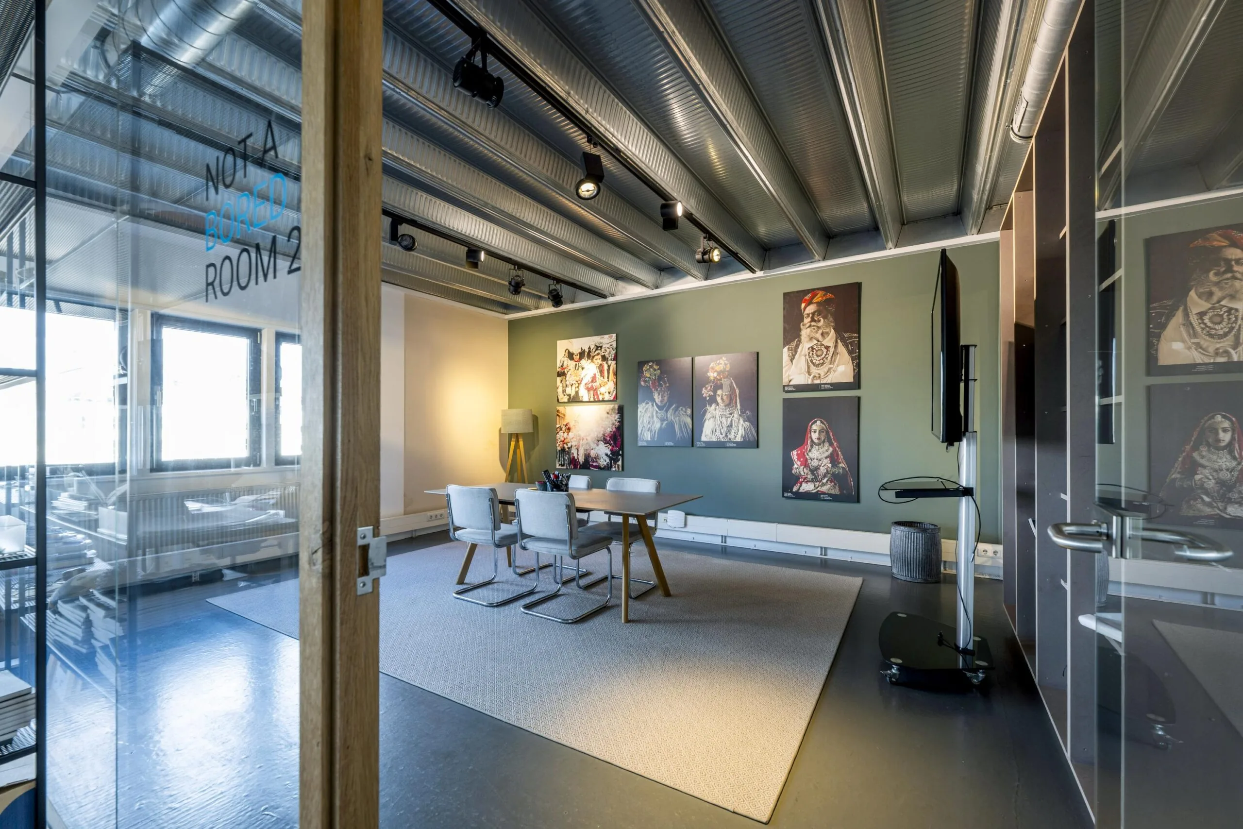 Modern meeting room at Helicopterstraat with industrial ceiling, art-covered wall, and a sign on the glass door reading "Not a Bored Room 2".