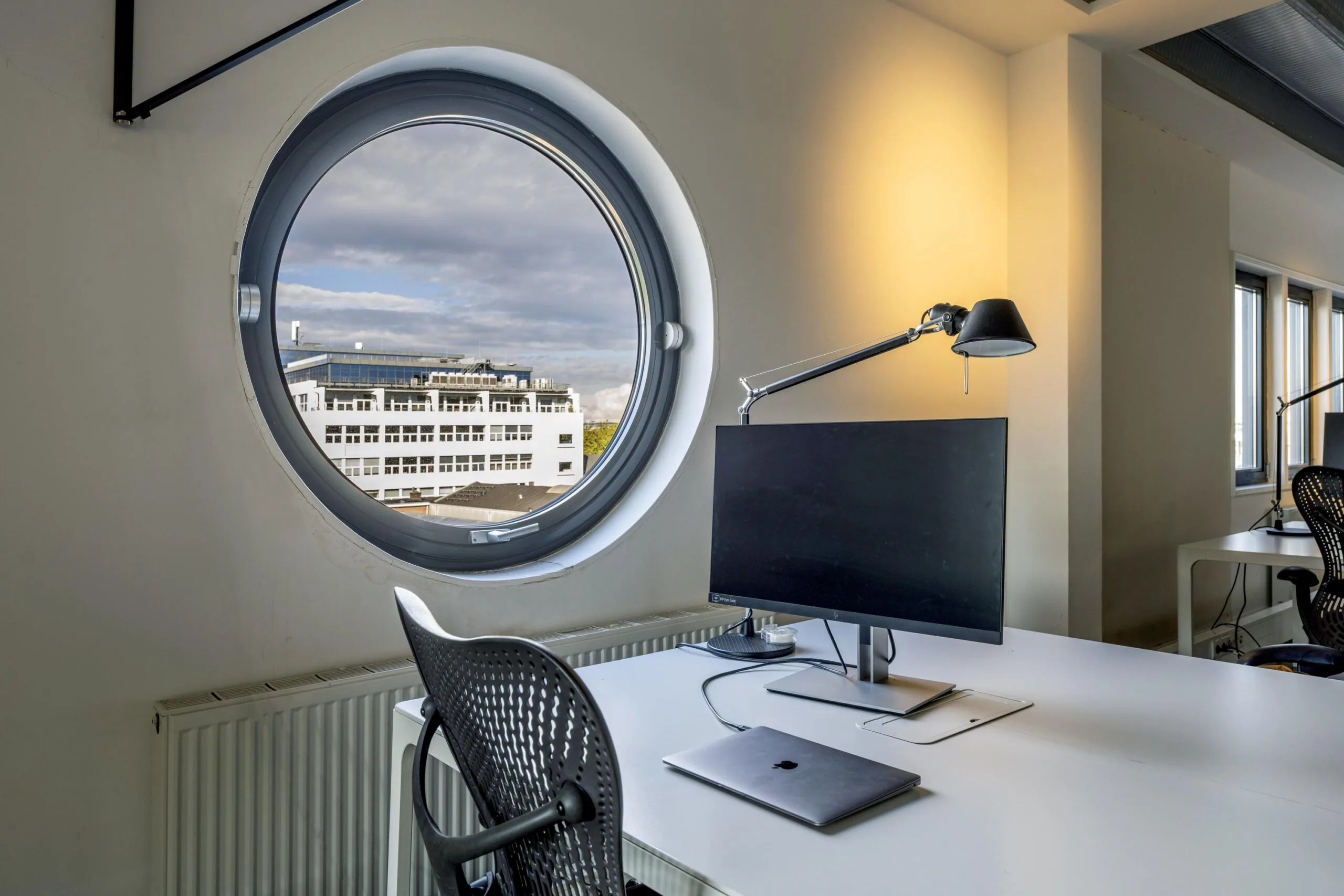 Modern office desk setup with a monitor and laptop near a round window overlooking a white building on Helicopterstraat.