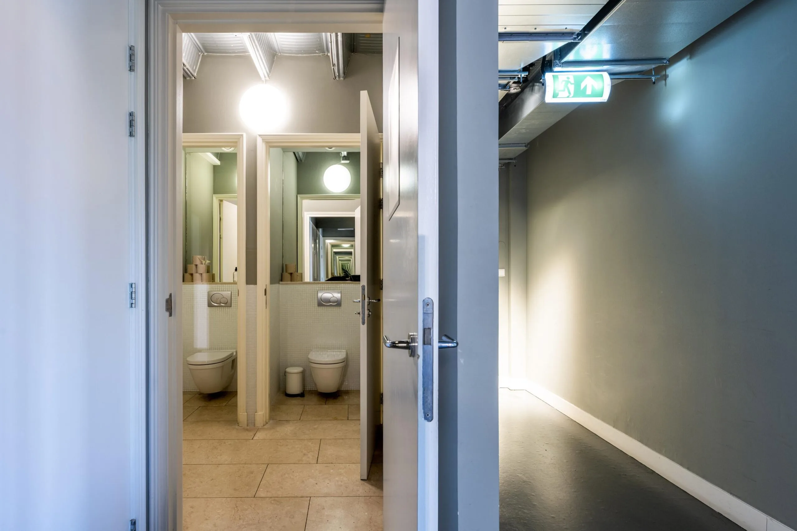Open door revealing a modern public restroom with mirrored reflections and an illuminated emergency exit sign above a hallway.