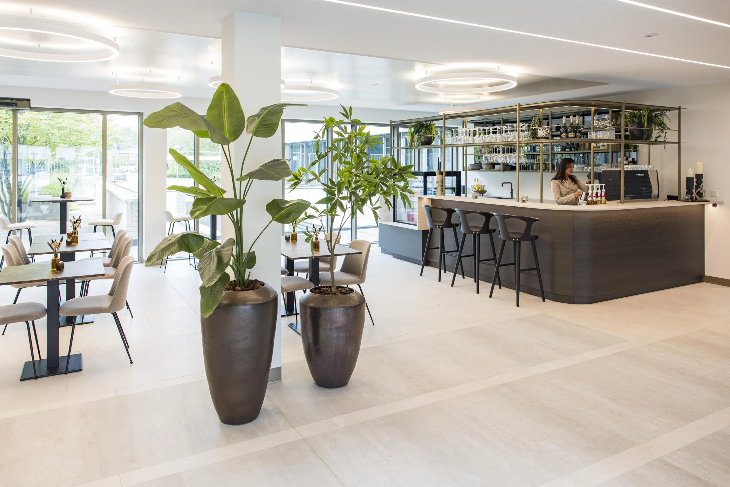 Modern hotel bar and seating area with large potted plants, minimalist furniture, and a woman preparing drinks behind the counter.