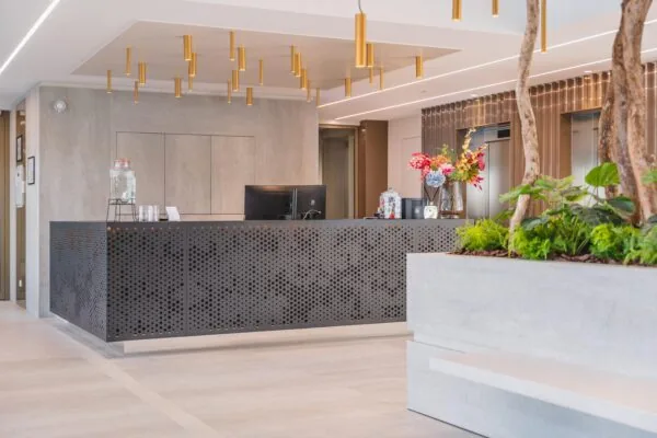 Modern reception desk in a bright office lobby with gold ceiling lights, fresh flowers, and indoor plants.