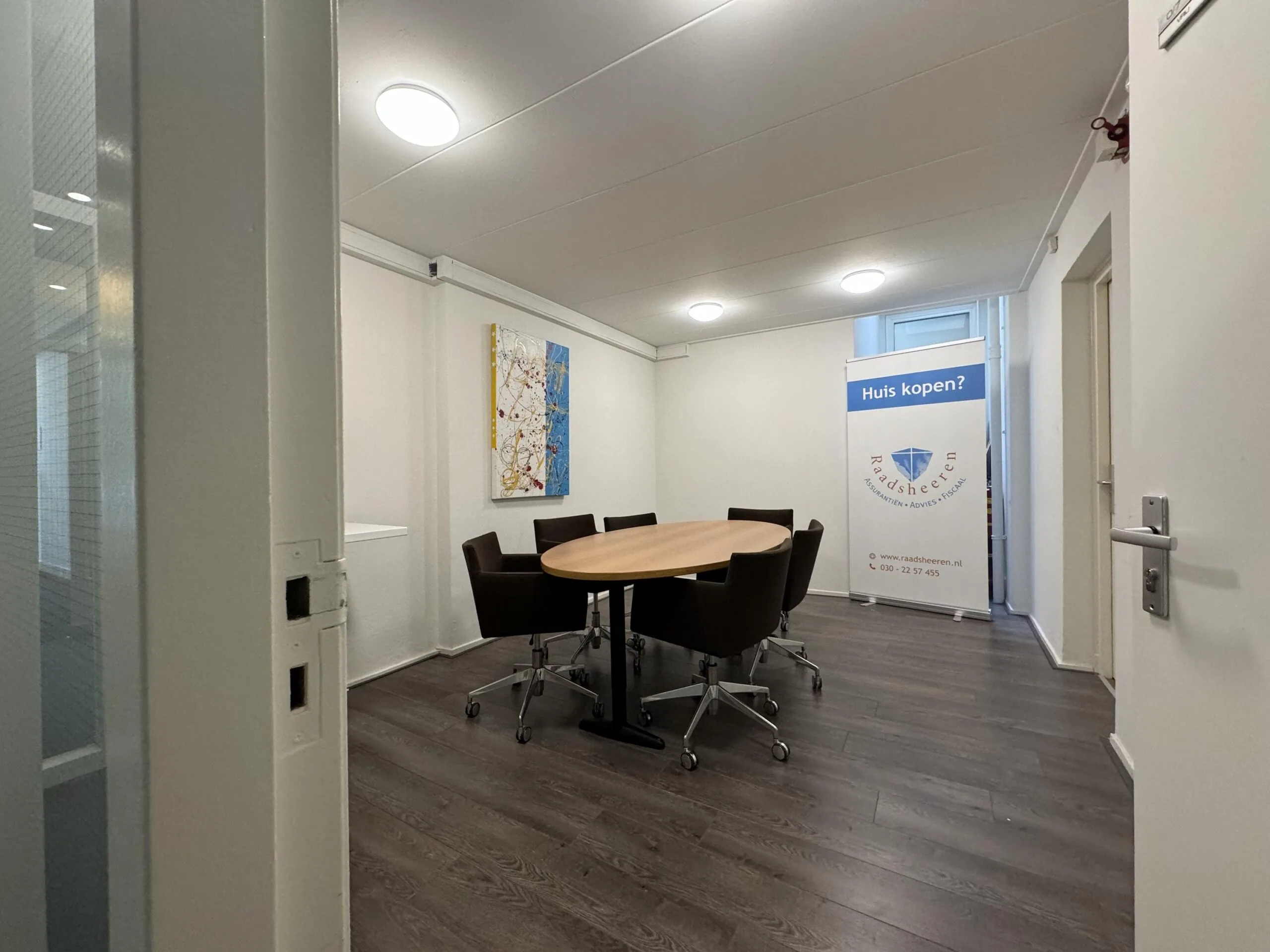 Modern meeting room with a round table, office chairs, abstract wall art, and a banner advertising "Raadsheren" real estate services.