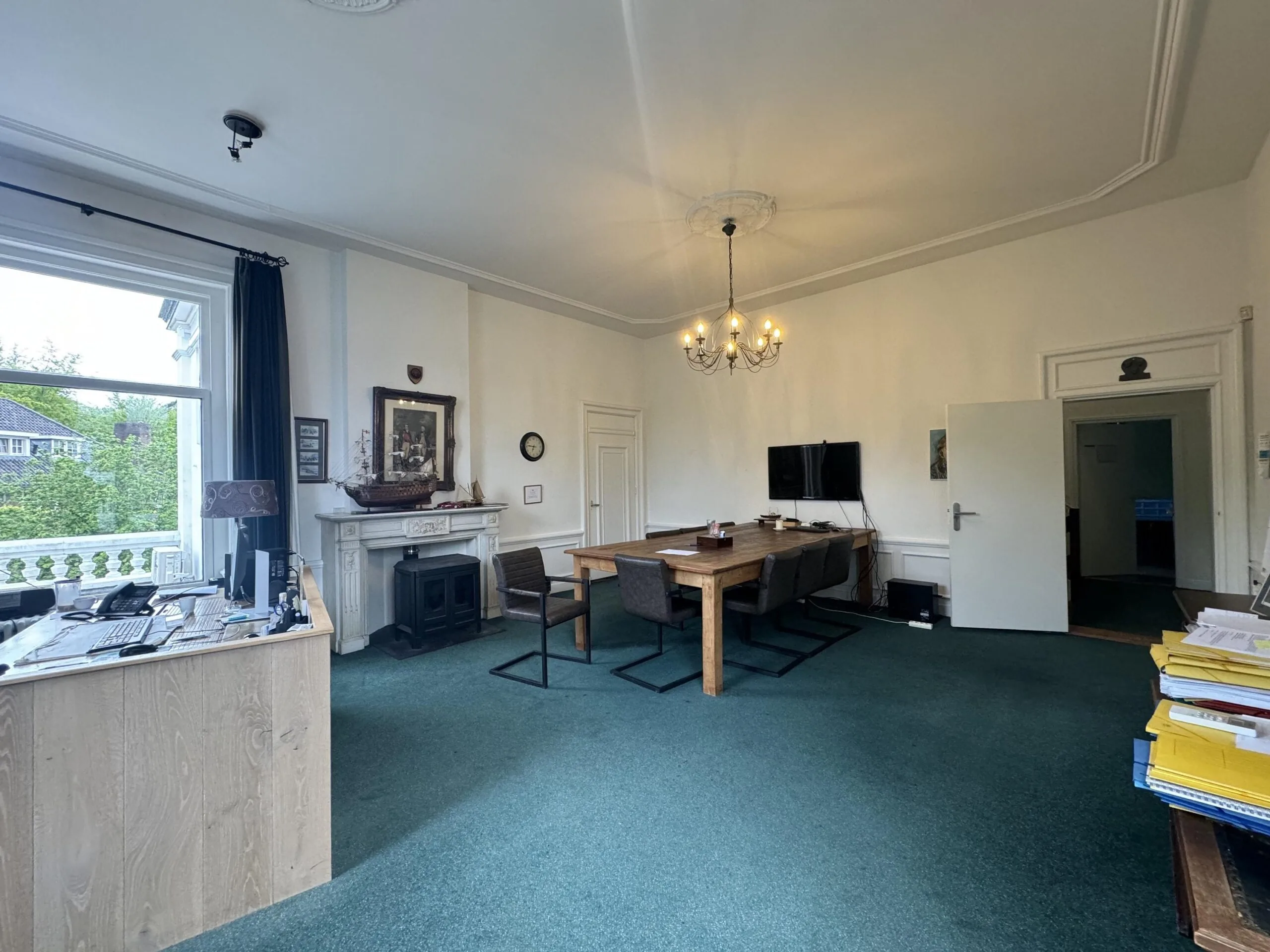 A spacious and traditional office room on Dorpsstraat near Steenstraat, featuring a large wooden table, fireplace, chandelier, and desk by the window.
