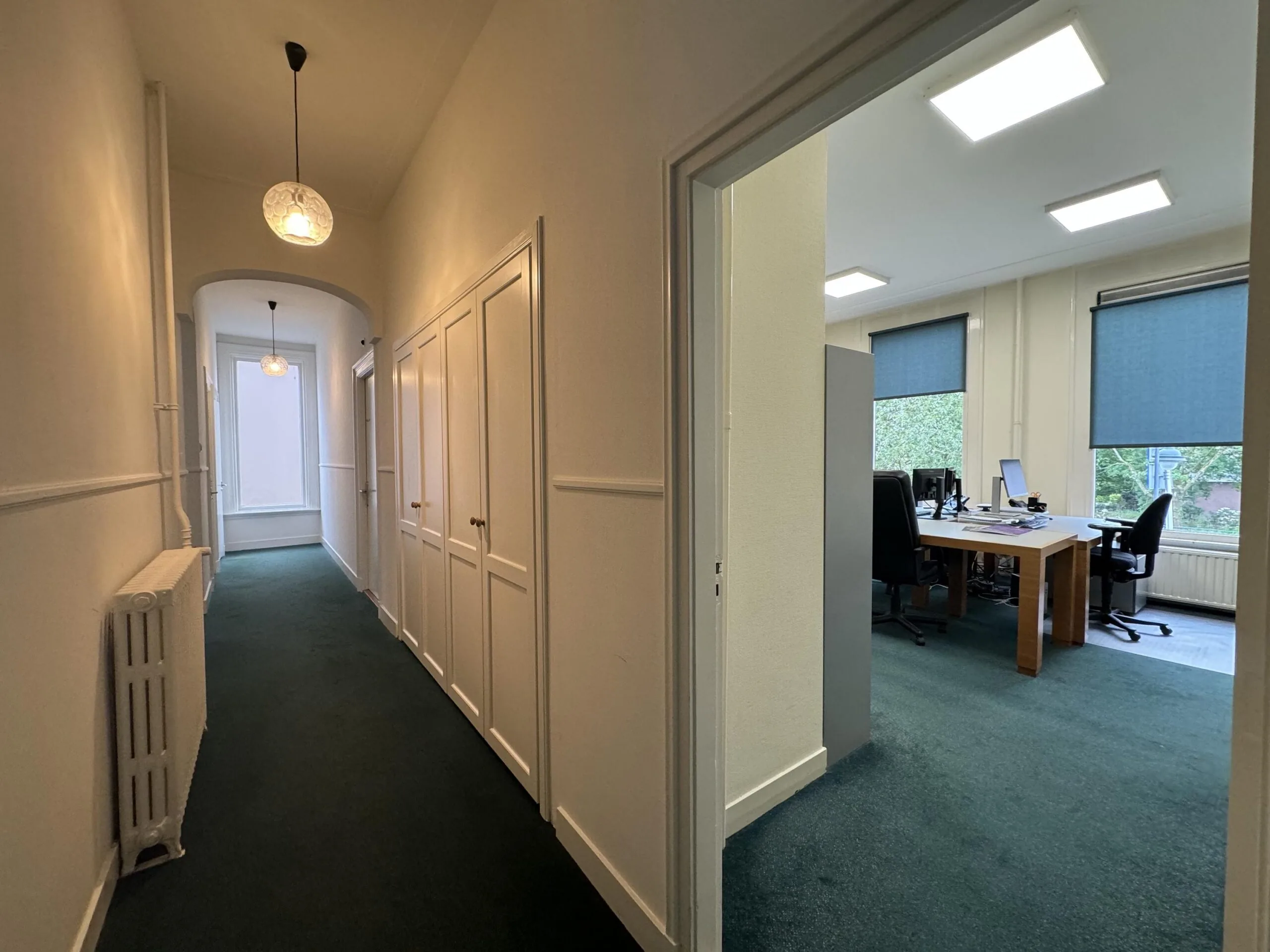 A hallway in an office building on Dorpsstraat facing Steenstraat, with a view into a workspace containing desks and office chairs.