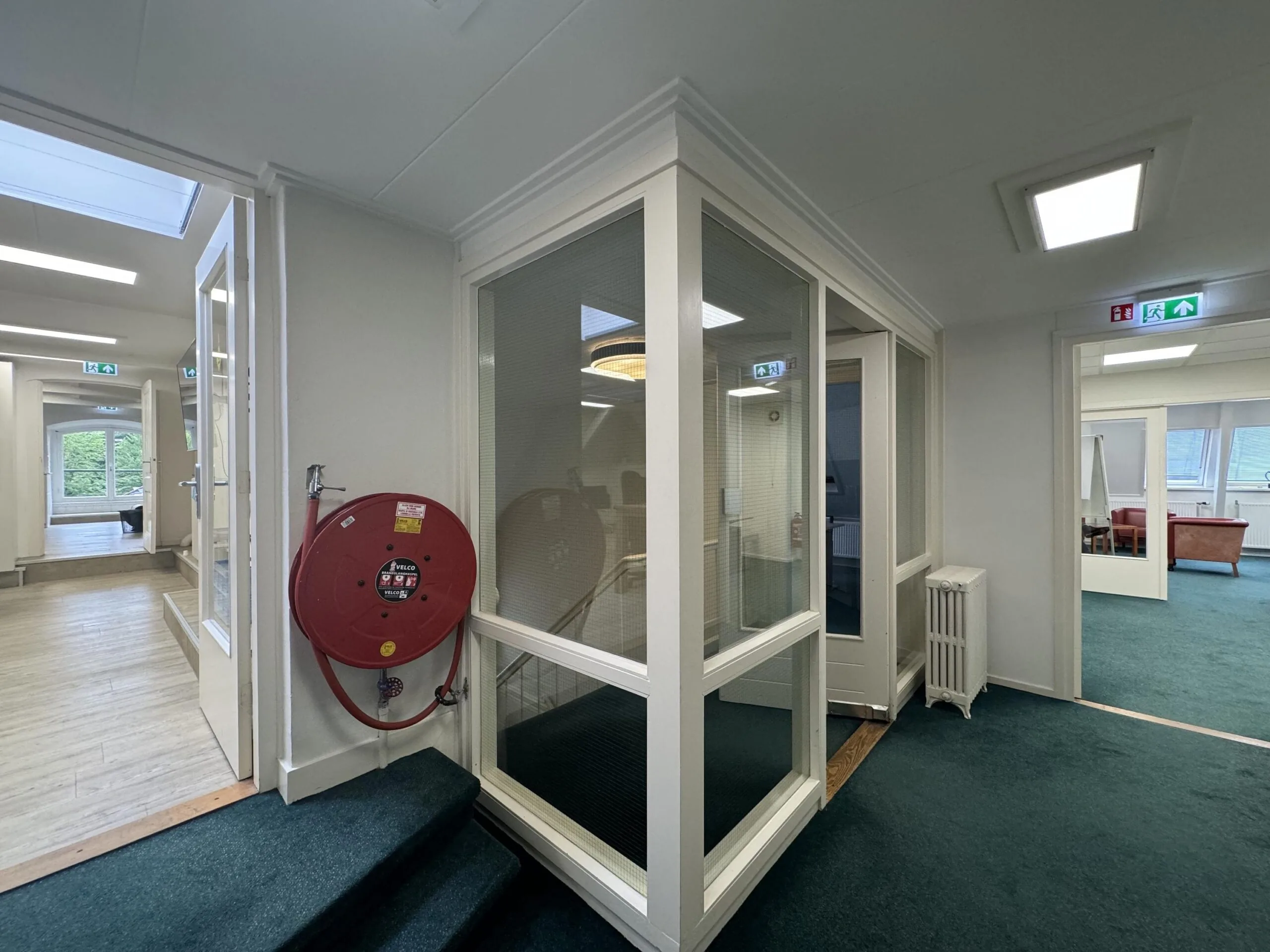 Office hallway at Dorpsstraat near Steenstraat with green carpet, a fire hose reel, and multiple rooms and exit signs visible.