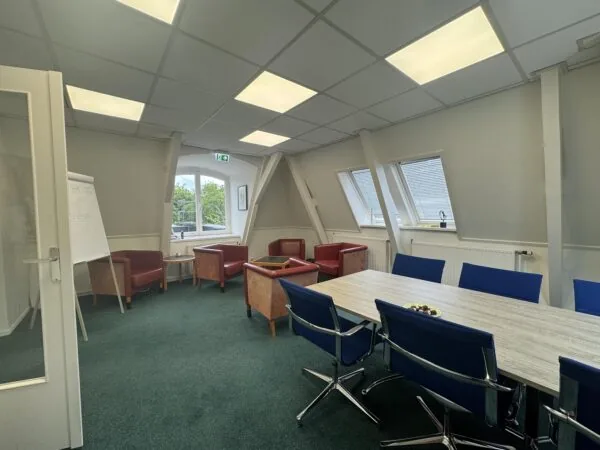 Meeting room with a long table, blue chairs, lounge corner with armchairs, and a whiteboard near the window on Dorpsstraat before Steenstraat.