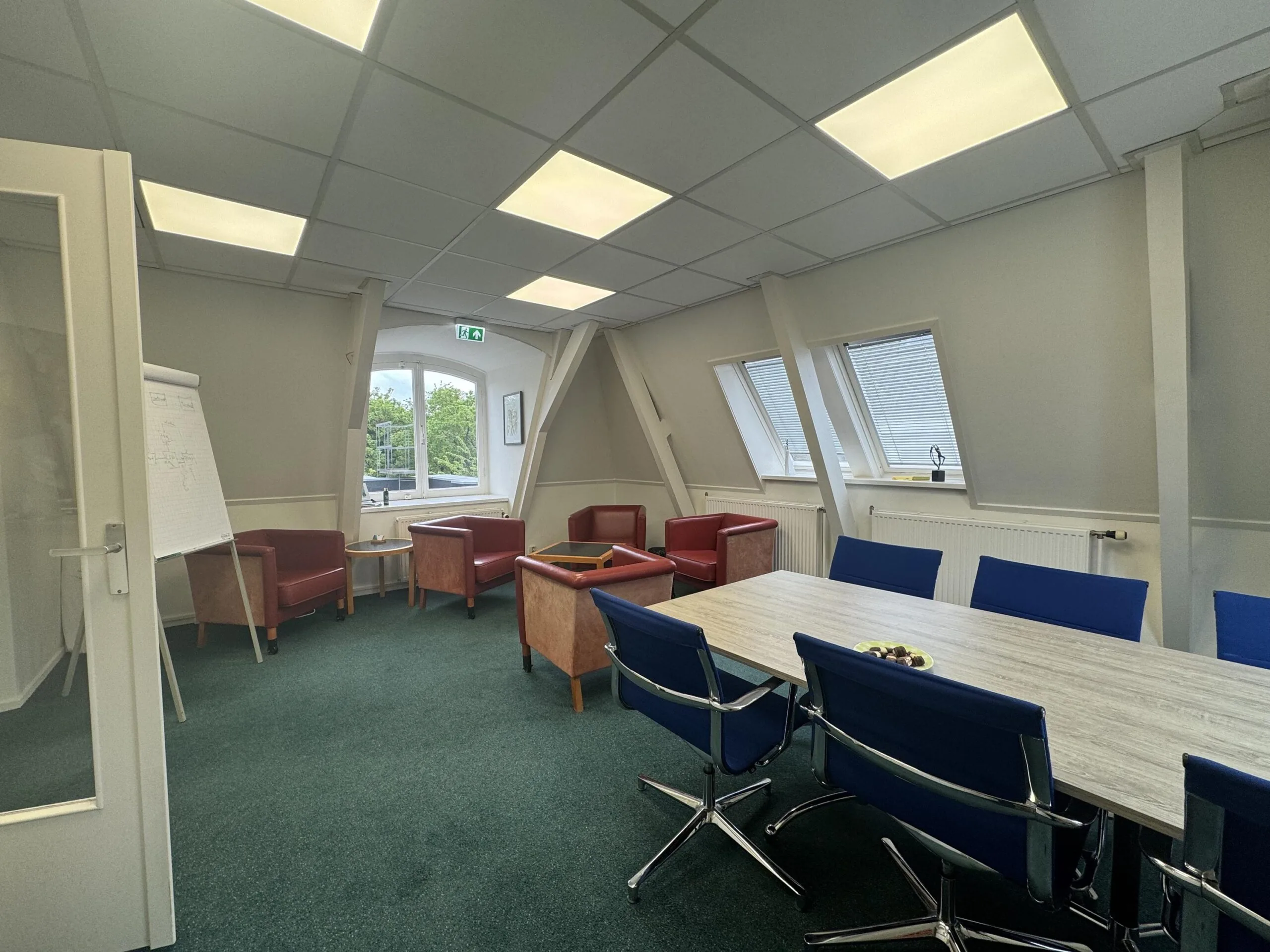 Meeting room with a long table, blue chairs, lounge corner with armchairs, and a whiteboard near the window on Dorpsstraat before Steenstraat.