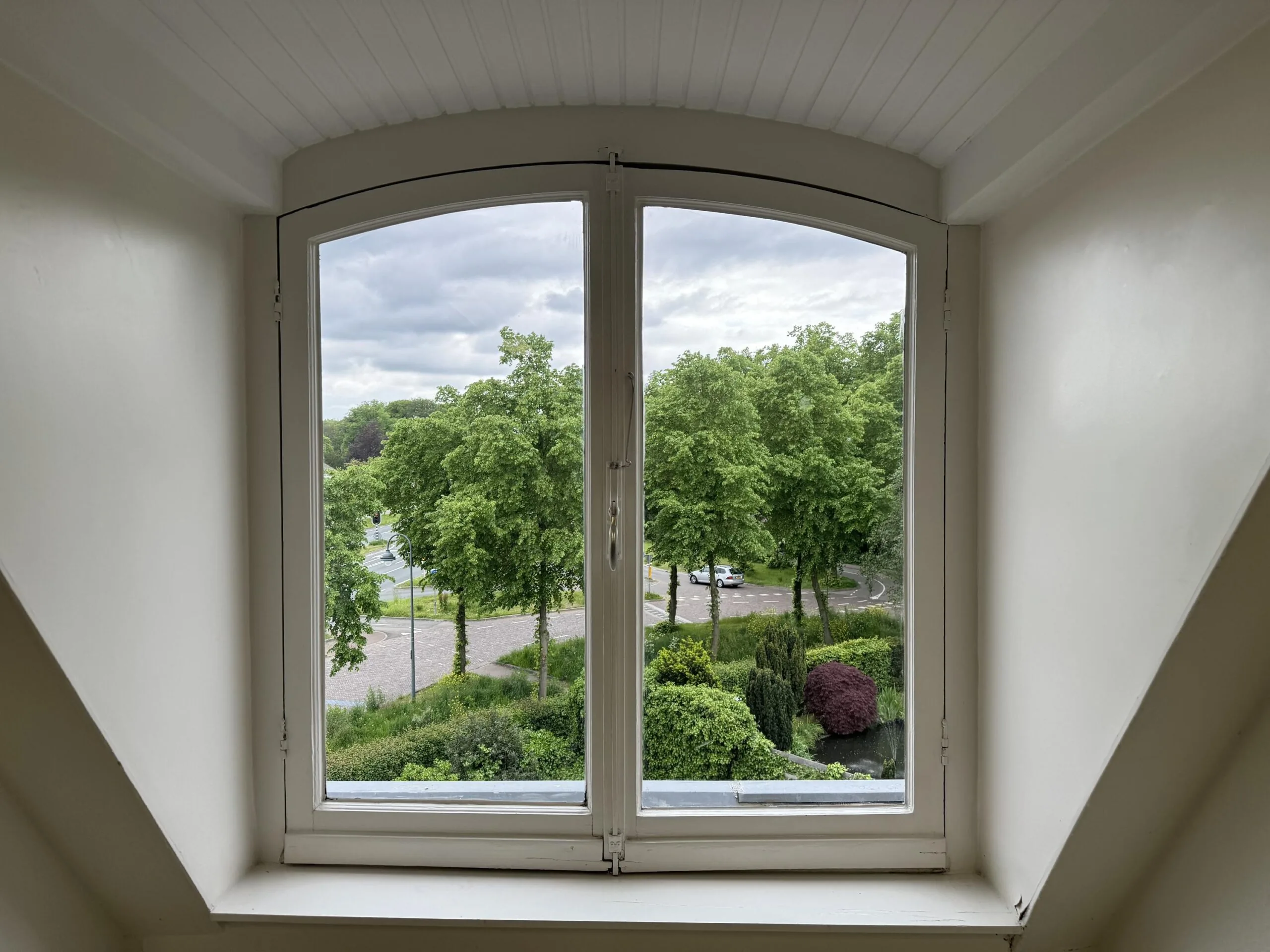 View through an arched attic window overlooking Dorpsstraat and Steenstraat, with trees, parked cars, and greenery visible outside.