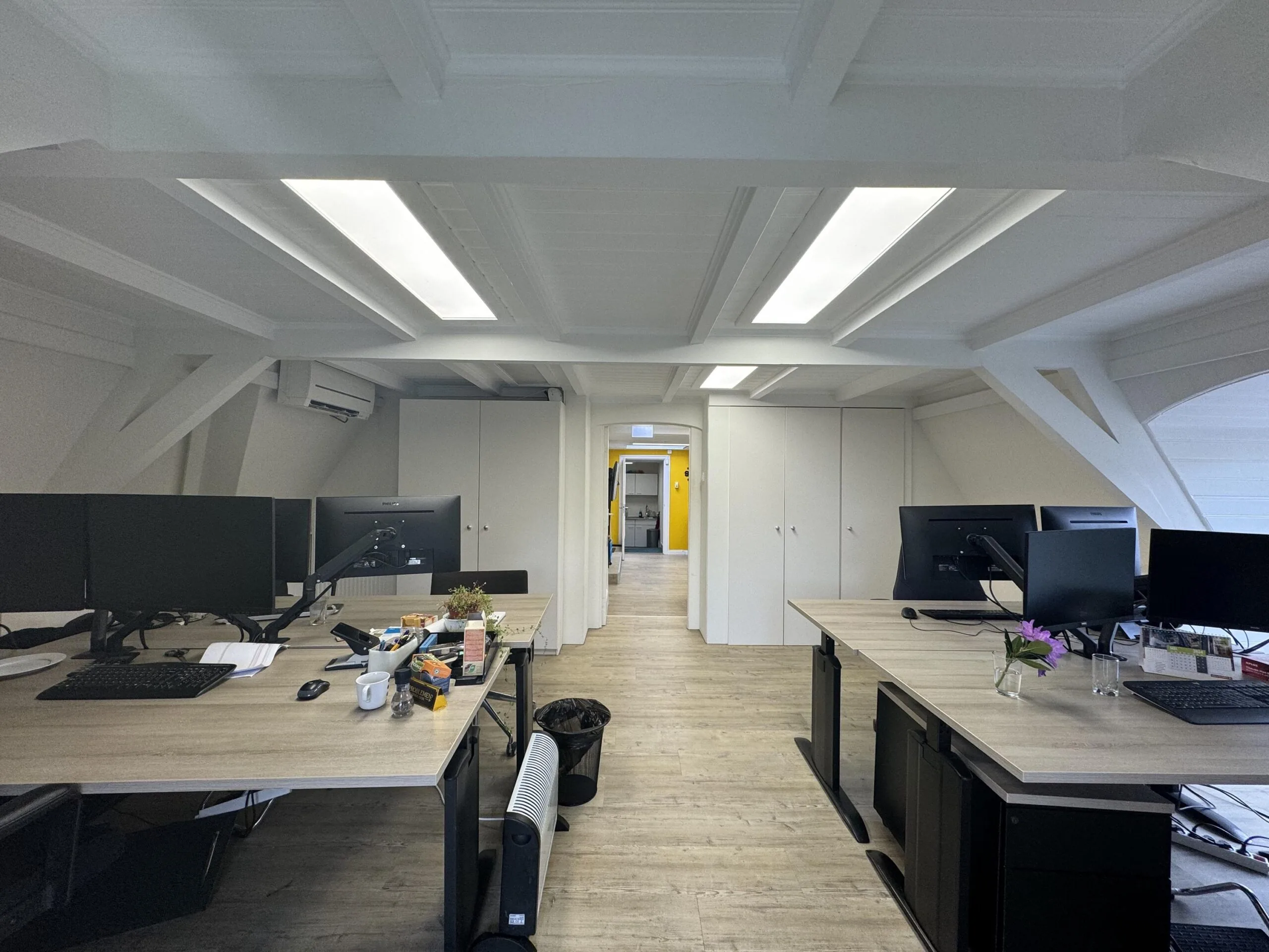 Modern open-plan office space with wooden desks, computer monitors, and a hallway with a yellow accent wall in the background.