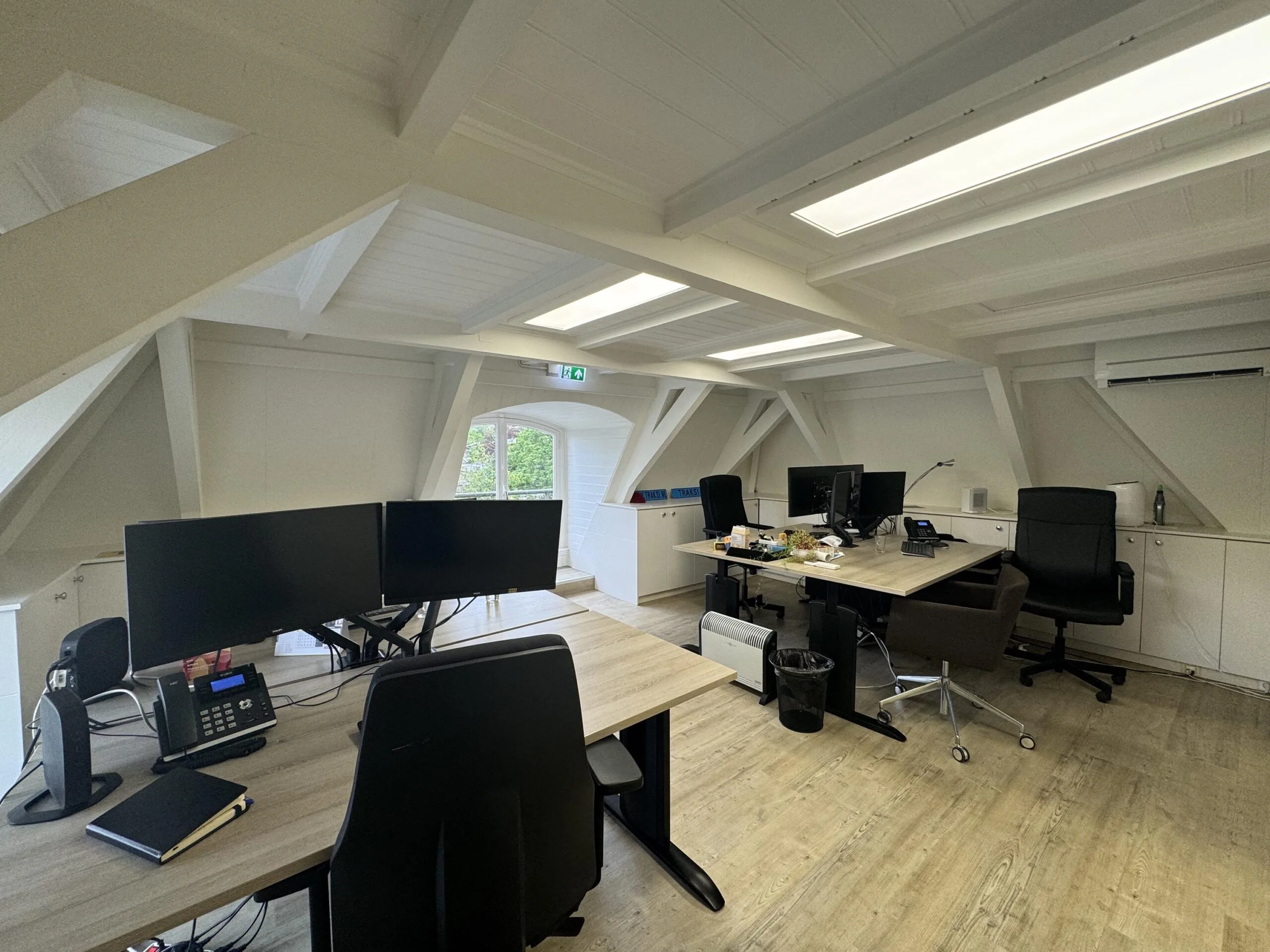 Modern attic office on Dorpsstraat before the intersection with Steenstraat, featuring multiple desks, monitors, and office chairs under a white wooden ceiling.