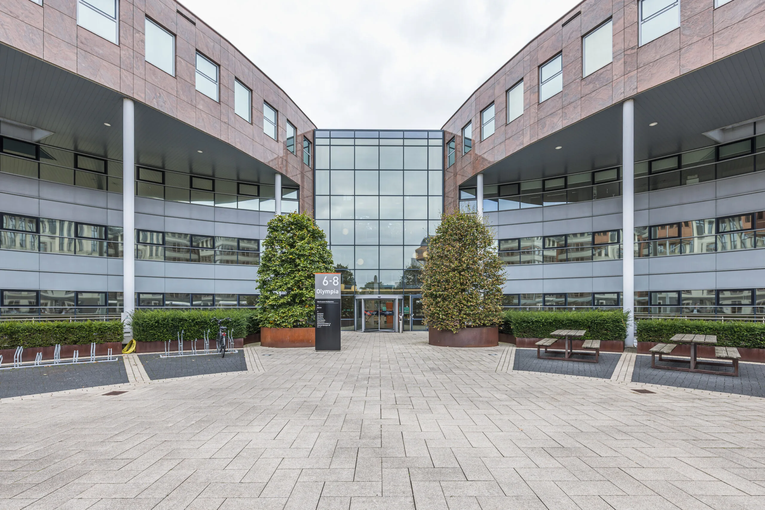 Modern office building named Olympia located at number 6-8, with a glass entrance, symmetrical architecture, and outdoor seating and bike racks.