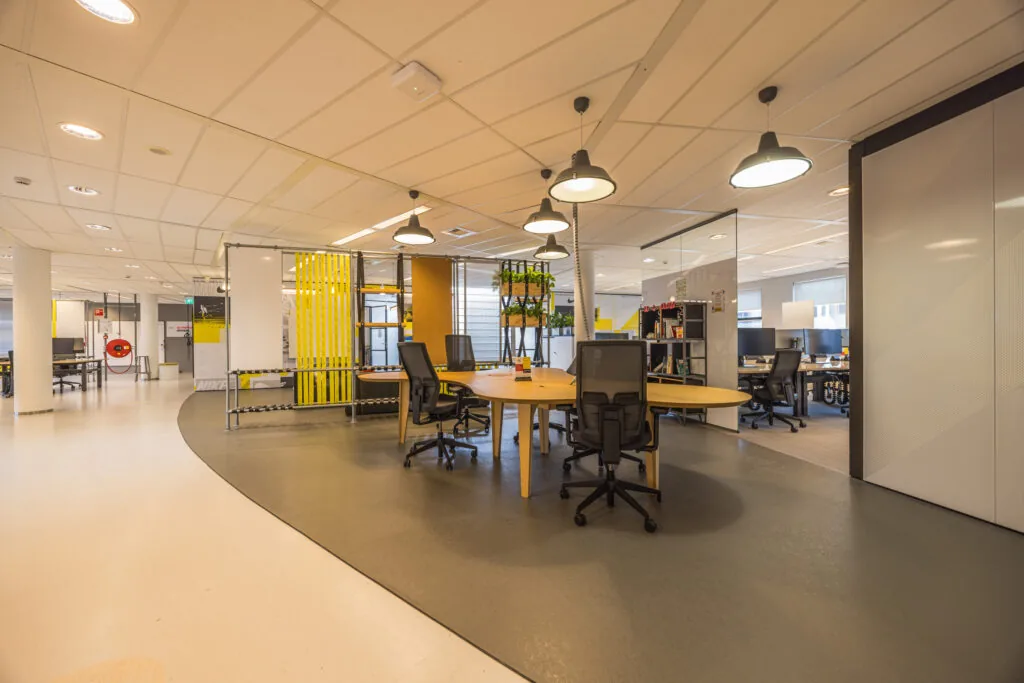 Modern open-plan office space with workstations, round tables, and bright yellow design accents.