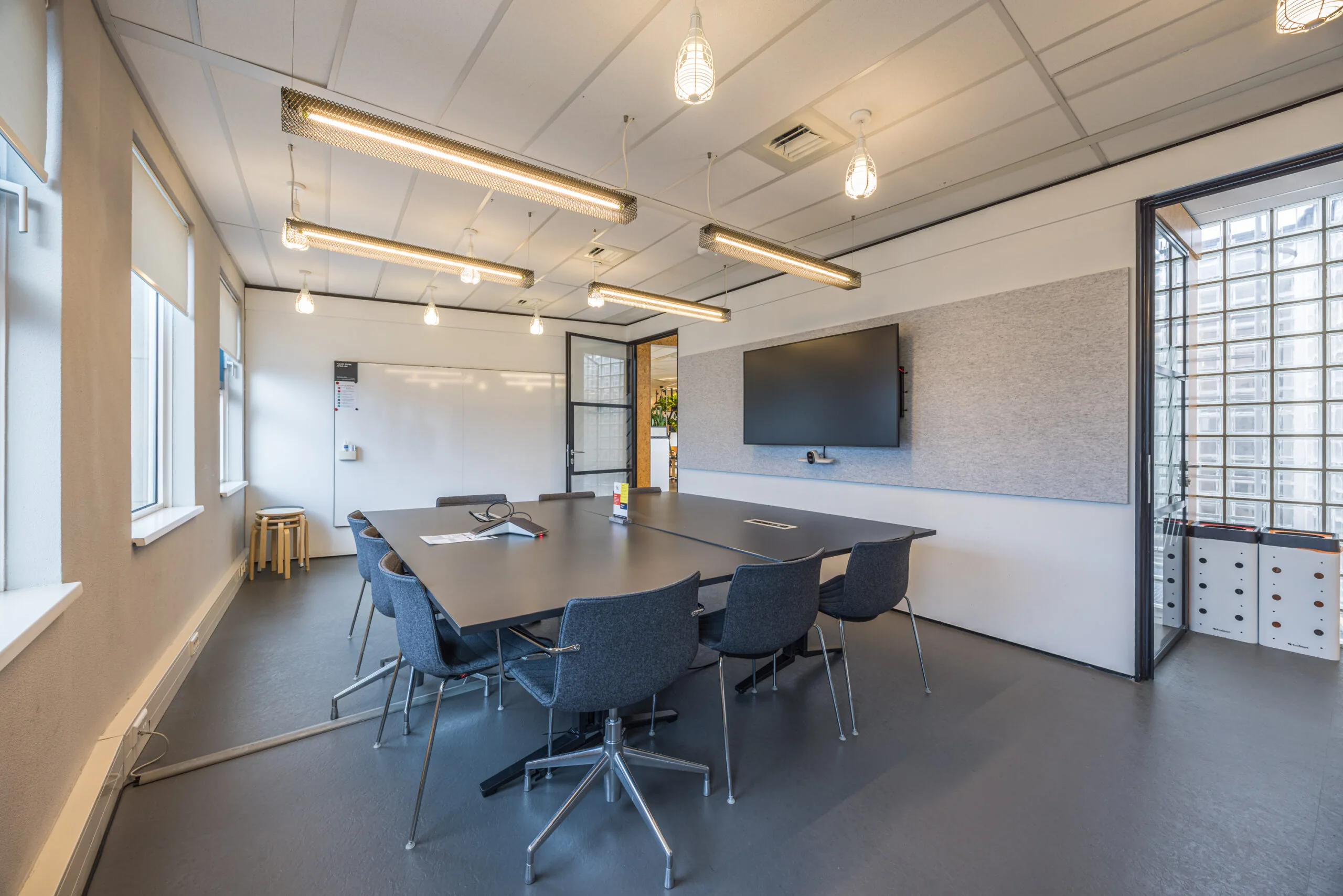 Modern conference room with a large meeting table, office chairs, a wall-mounted screen, and natural light from tall windows.