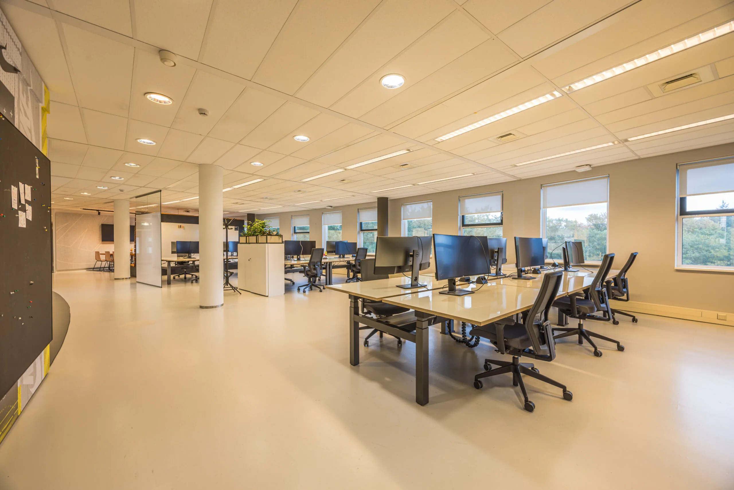 Modern open-plan office space with multiple desks, ergonomic chairs, and computer monitors under bright ceiling lights.