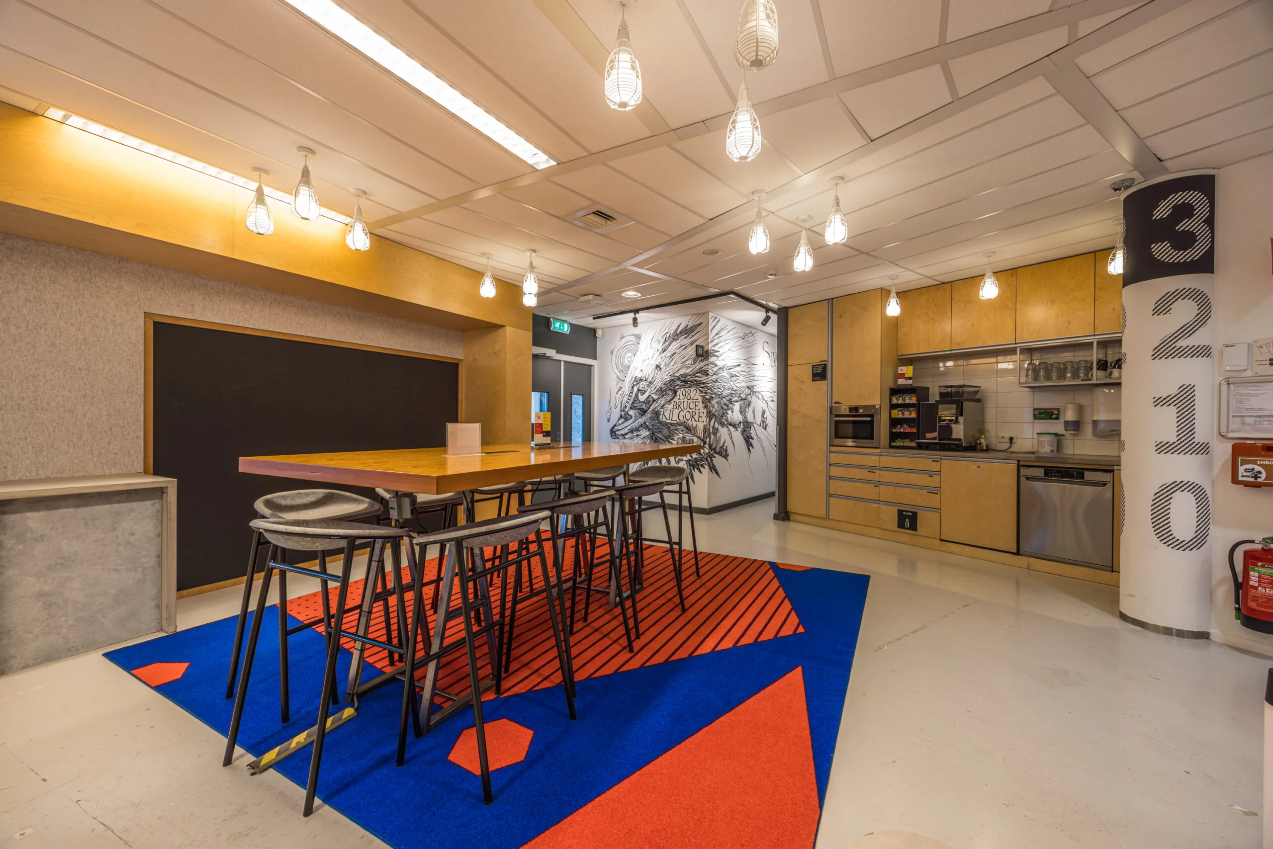 Modern office kitchen and lounge area with a high table, vibrant carpet, wall art, and built-in appliances.