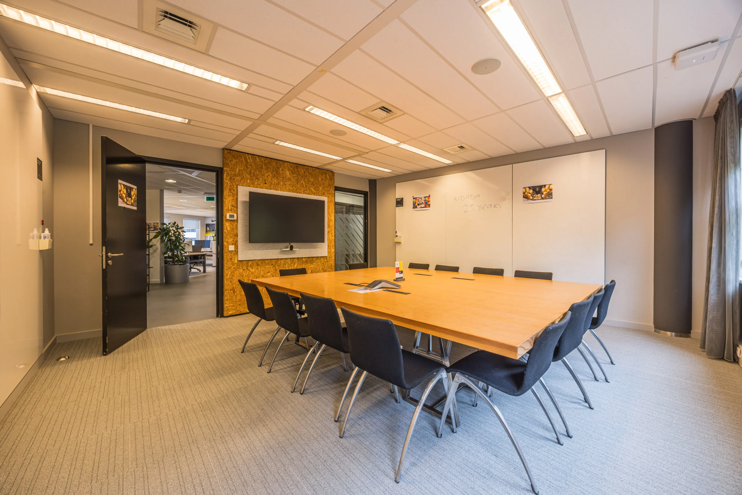 Modern conference room with a large wooden table, black chairs, a wall-mounted screen, and whiteboards.