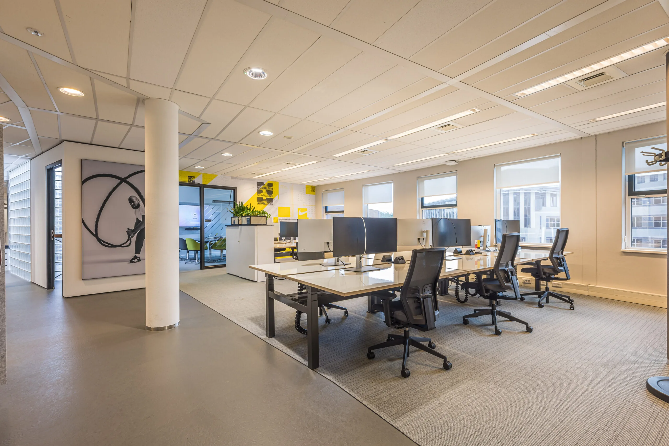 Modern open-plan office space with multiple workstations, ergonomic chairs, and a large wall portrait of a woman exercising with battle ropes.