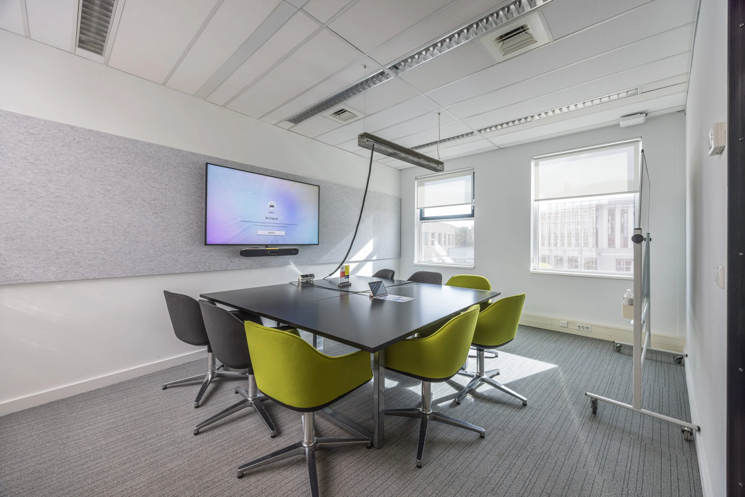 Modern conference room with a large screen, a black meeting table, green and gray chairs, and natural light from two windows.