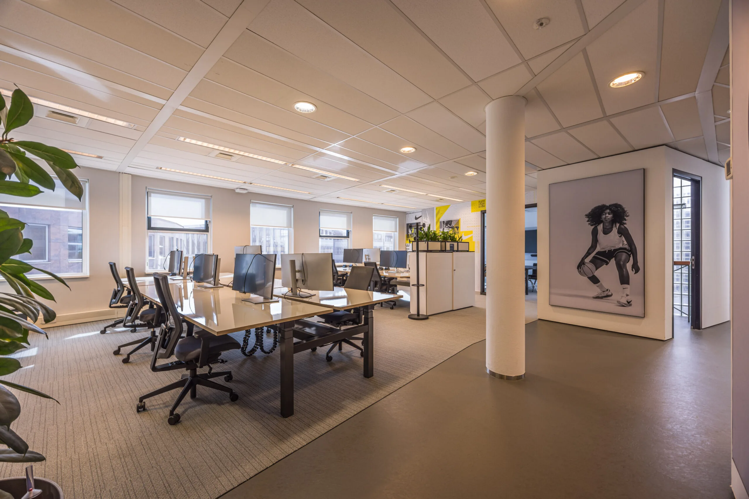 Modern open-plan office space with rows of desks, computer monitors, and a large black-and-white sports-themed wall poster.