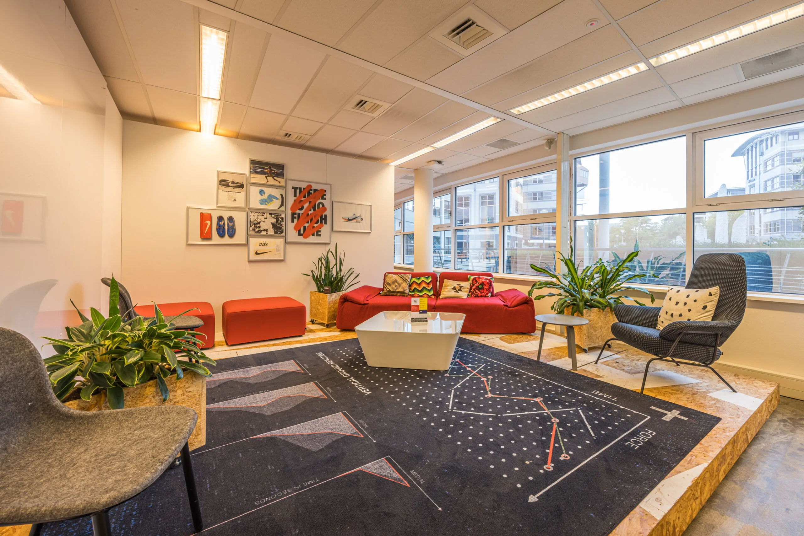 Modern office lounge area with colorful furniture, wall art, indoor plants, and large windows providing natural light.