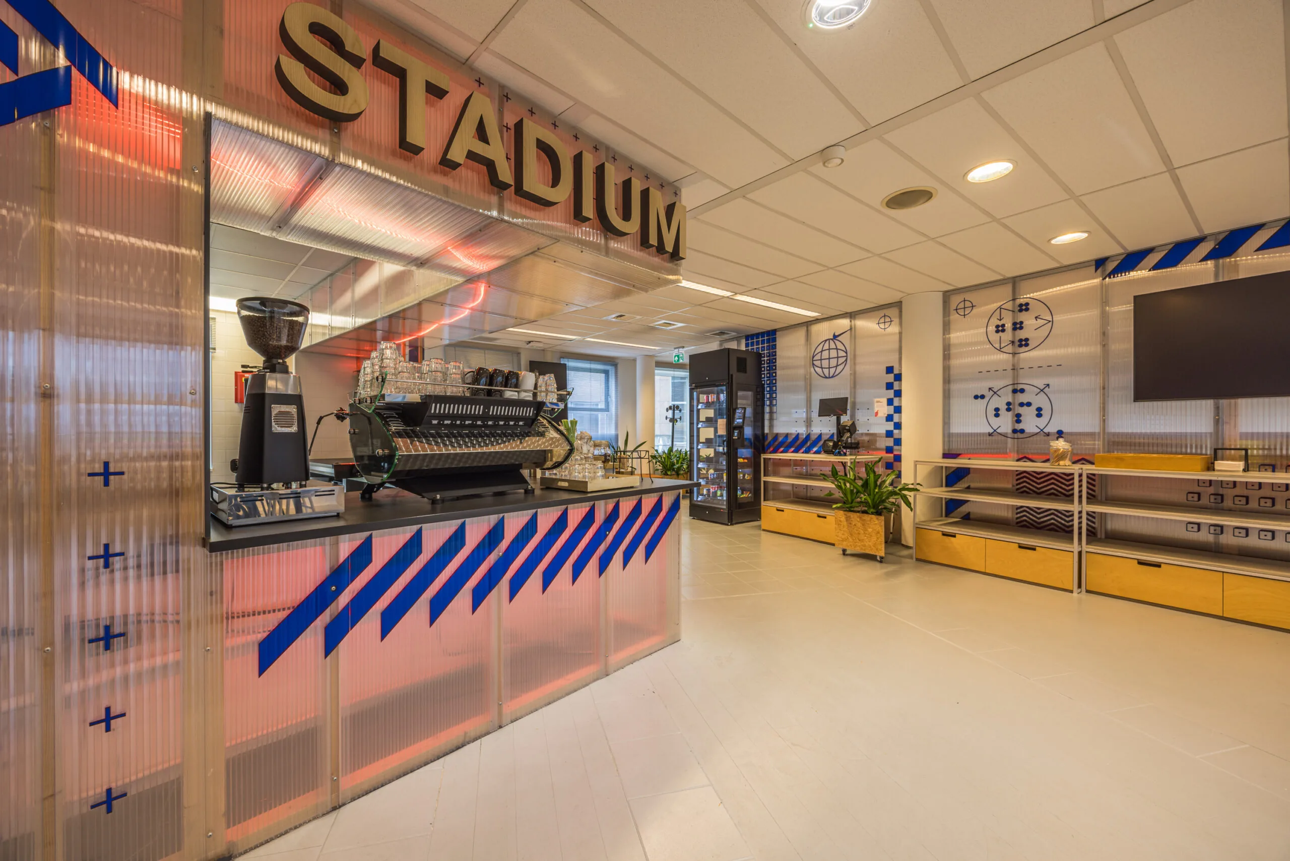 Modern coffee bar named "Stadium" with a sleek espresso machine and decorative blue-red graphic elements in an office-style interior.