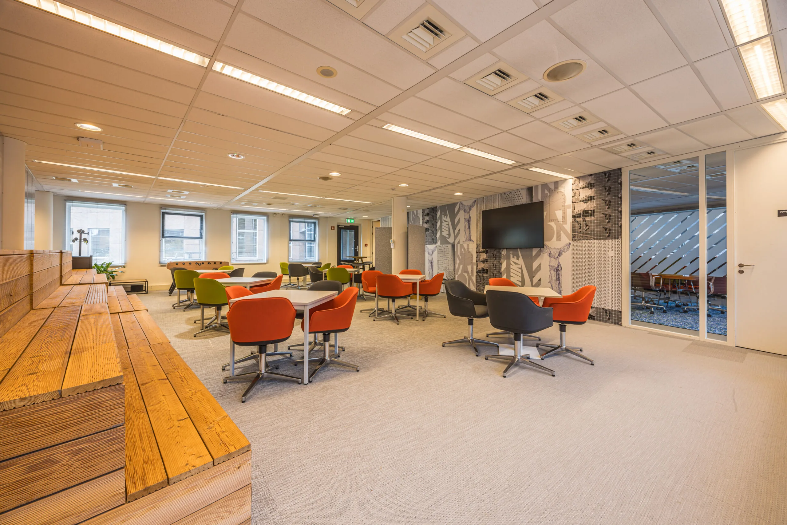 Modern office lounge area with colorful chairs, wooden seating steps, and a wall-mounted screen at Olympia.