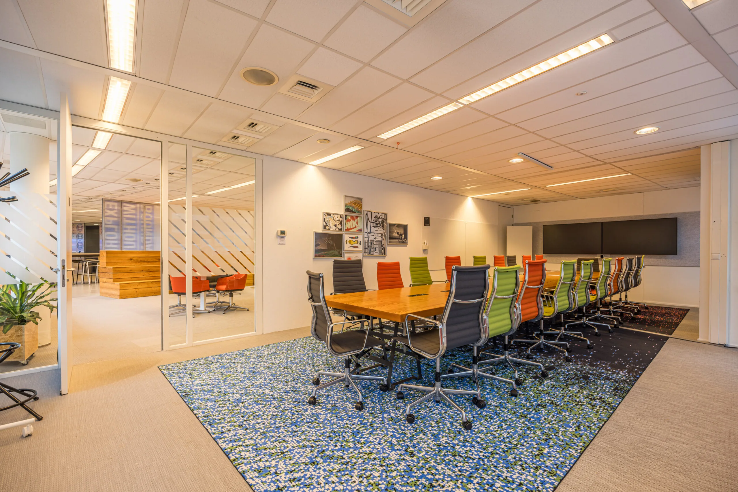 Modern conference room with a long wooden table, colorful office chairs, and large screens on the wall.