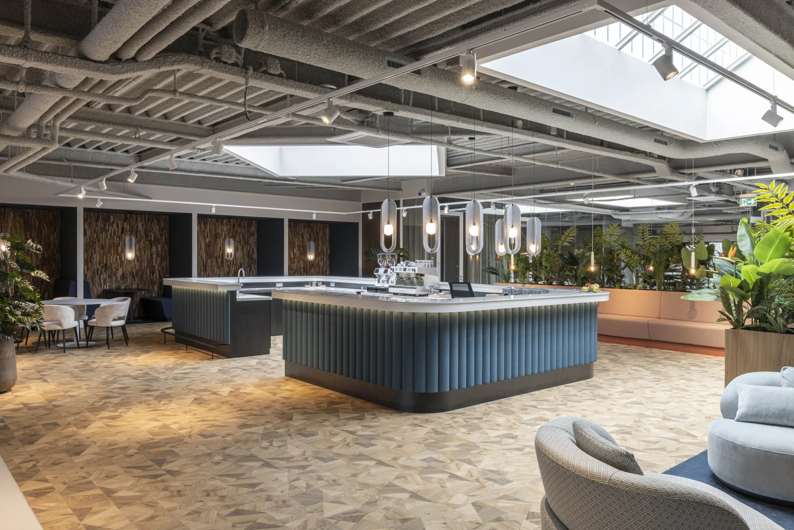 Modern office café area featuring a central bar with blue paneling, stylish lighting, and lush indoor plants at Essebaan.
