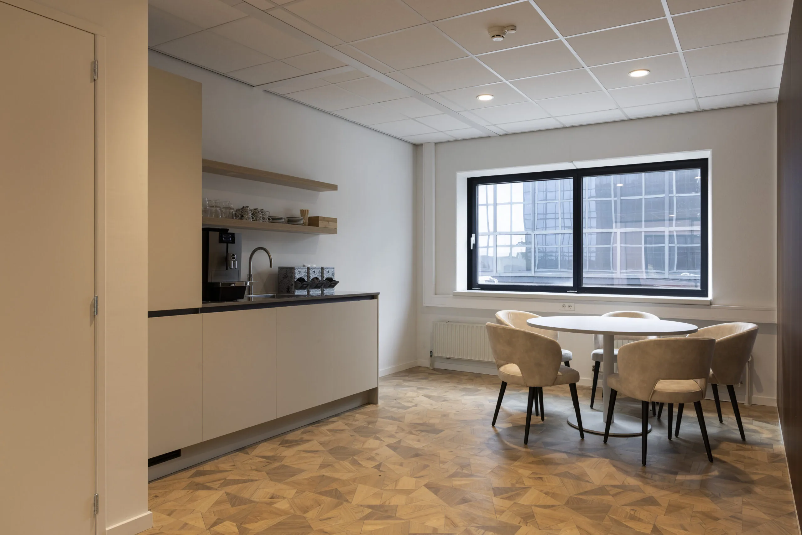 Modern office kitchenette with coffee machine, round table, and beige chairs next to a large window.