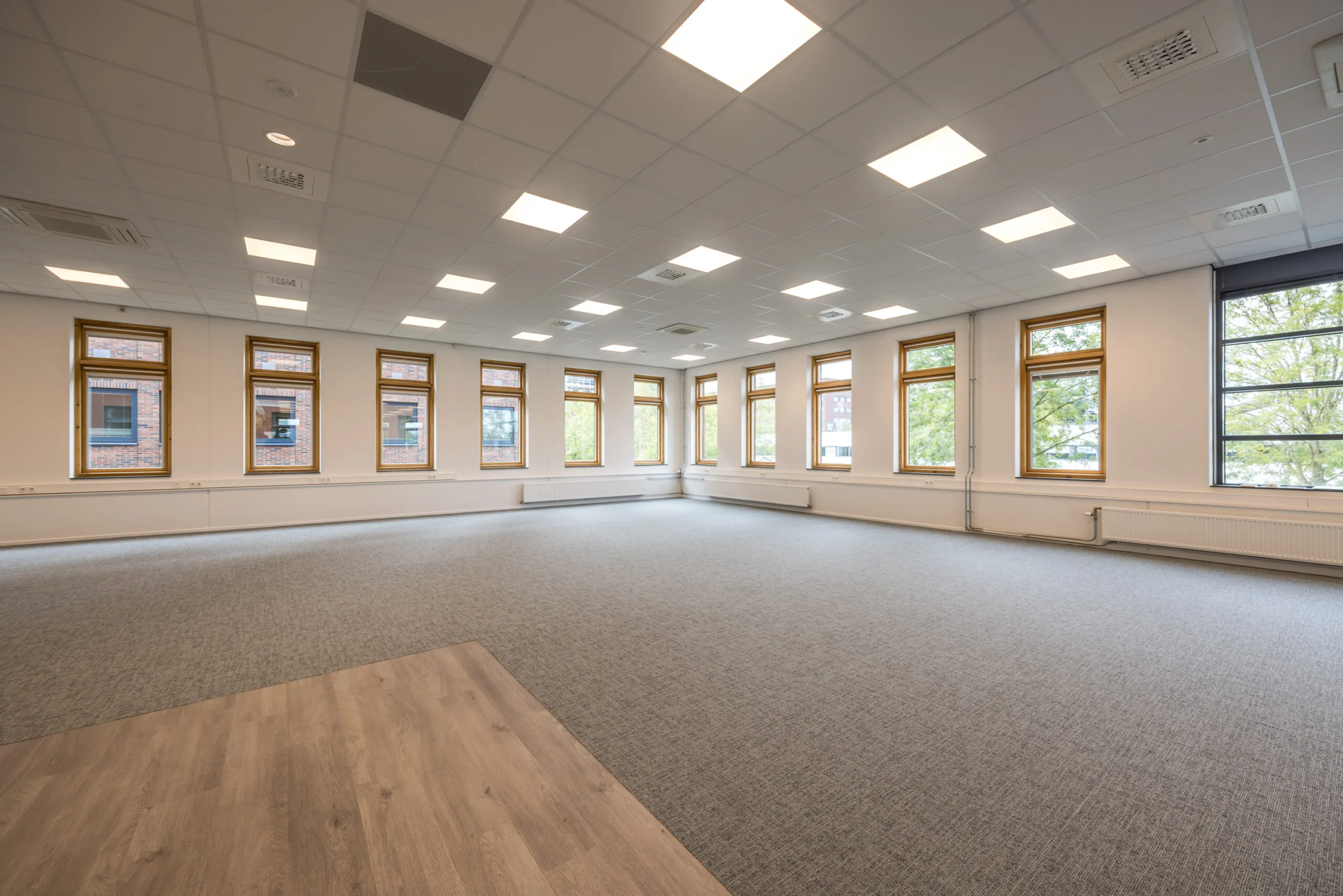 Spacious empty office space at Veldzigt with large windows, carpeted floor, and ceiling lighting.