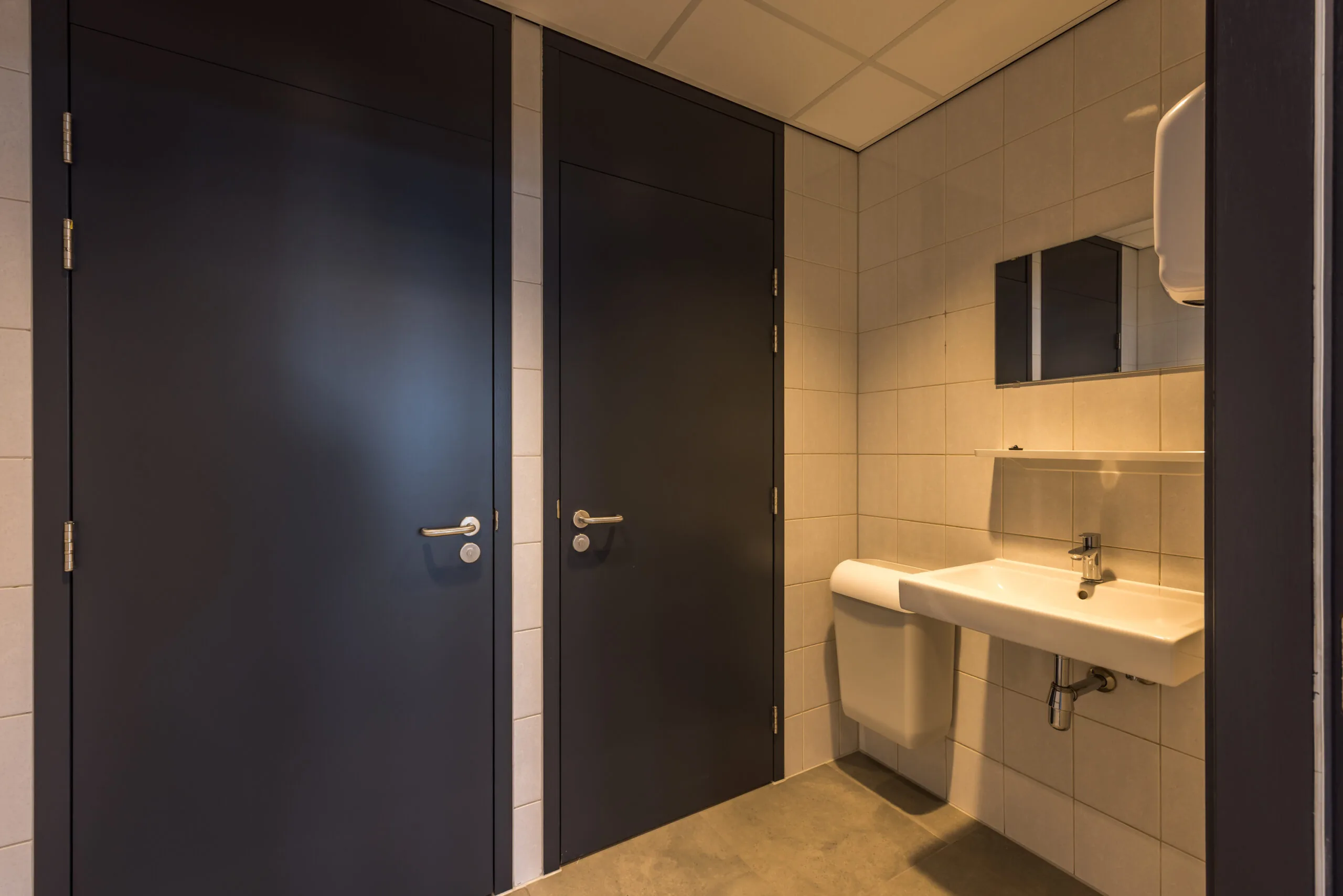 Modern public restroom with two dark-colored stall doors and a white sink beneath a mirror.