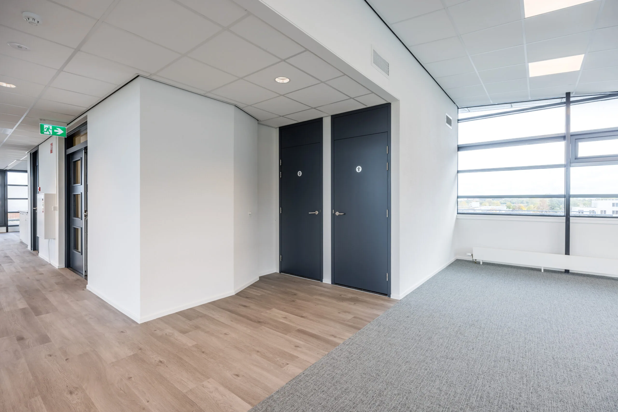 Modern office hallway with two dark restroom doors, wood flooring, and large windows providing natural light.