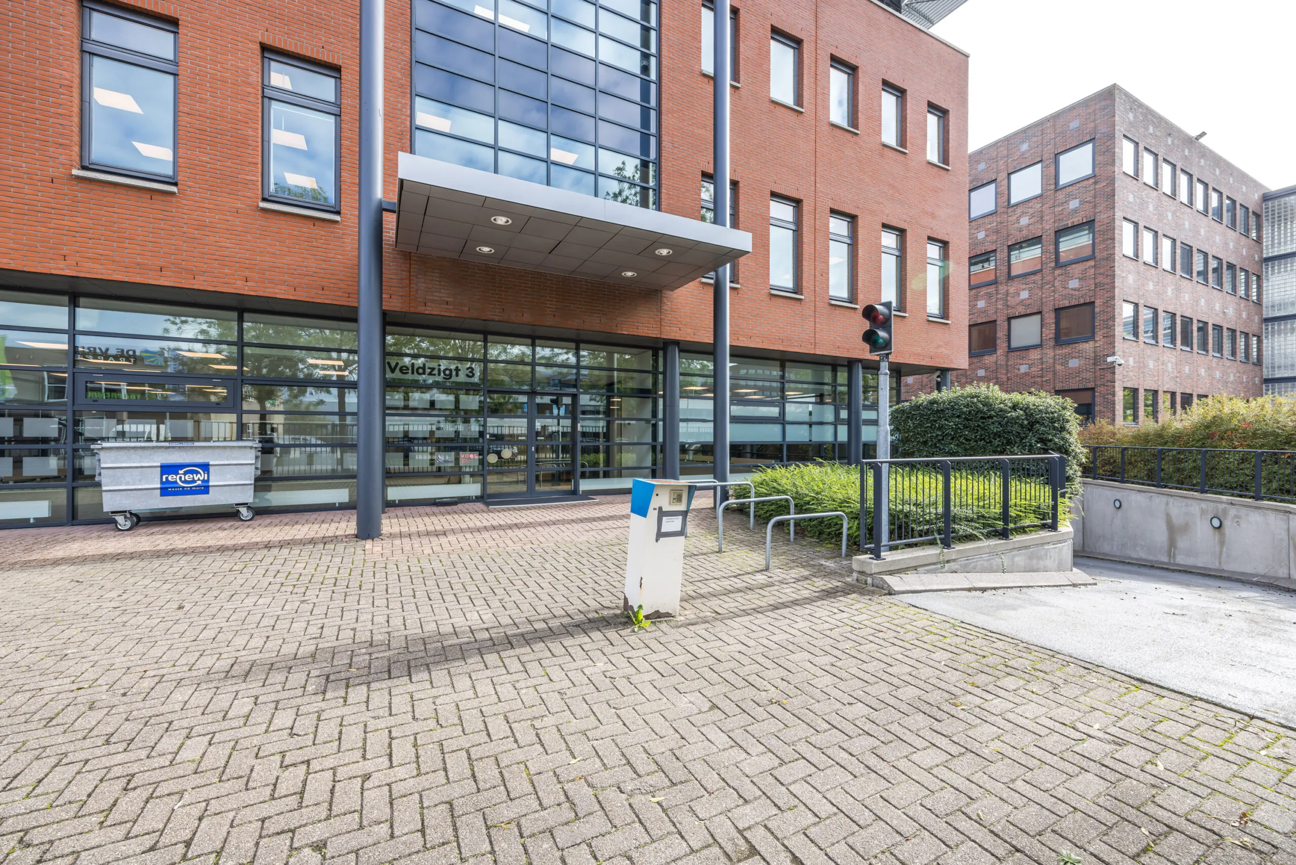 Modern office building at Veldzigt 3 with a glass entrance, a waste container, and an underground parking ramp.